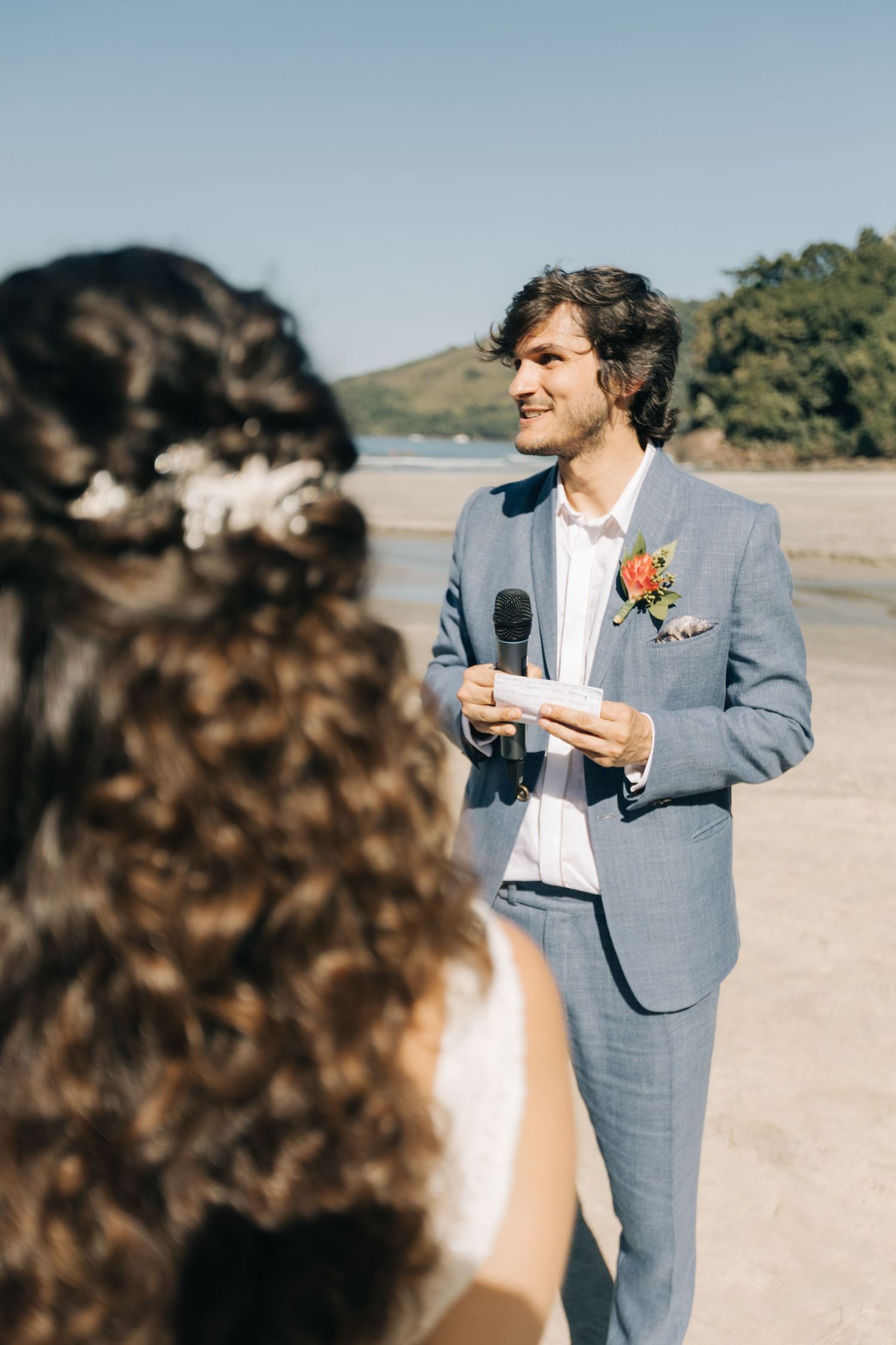 A wedding ceremony takes place on a scenic beach with mountains in the background.