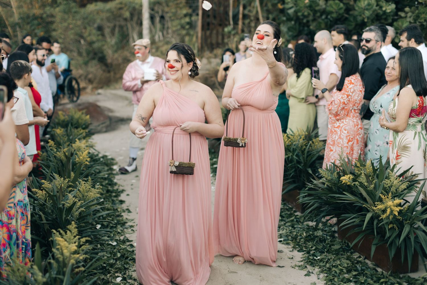 Bridesmaids in flowing pink gowns walk down a garden path during a wedding ceremony.