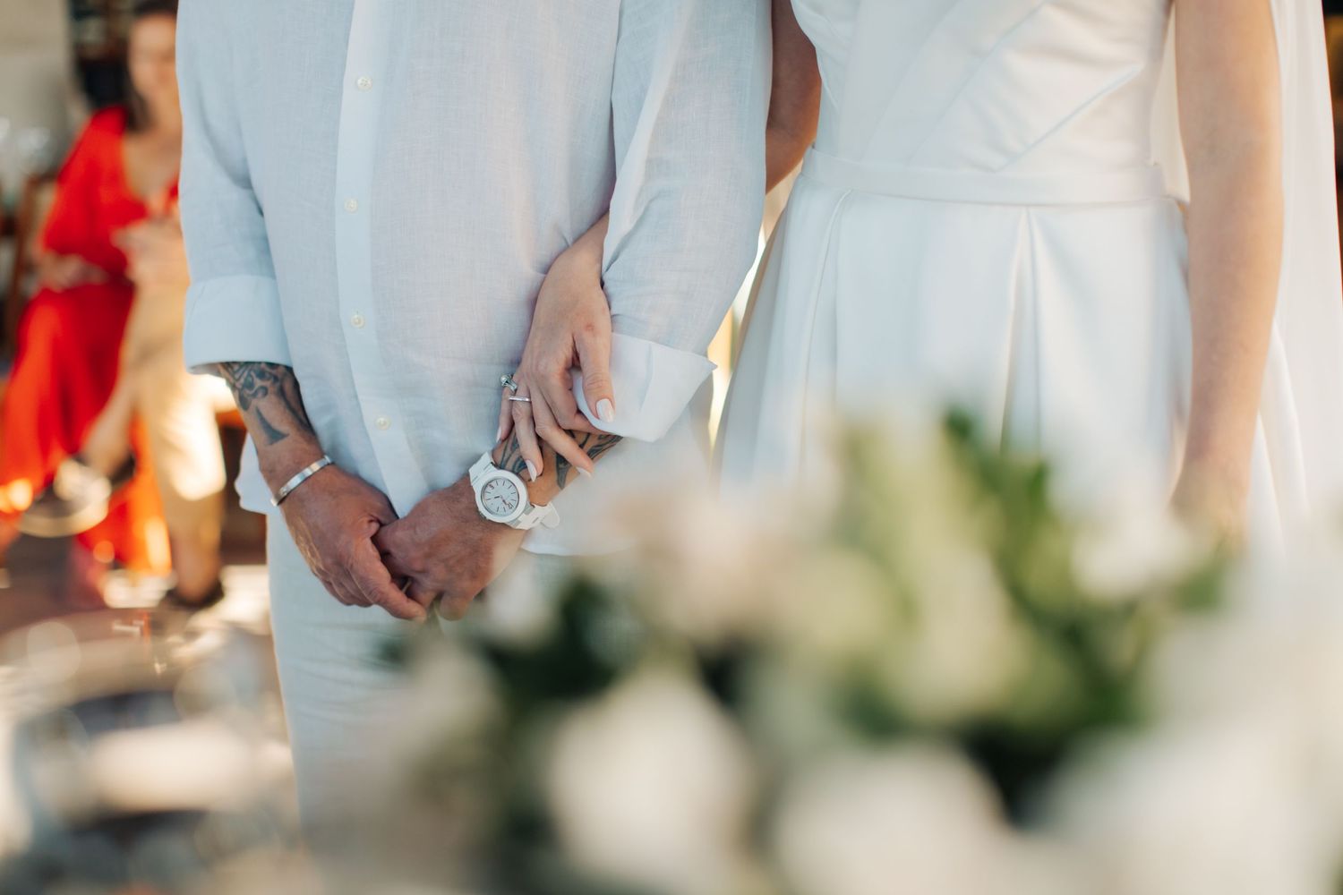 Close-up of clasped hands during wedding ceremony with floral decorations in background.