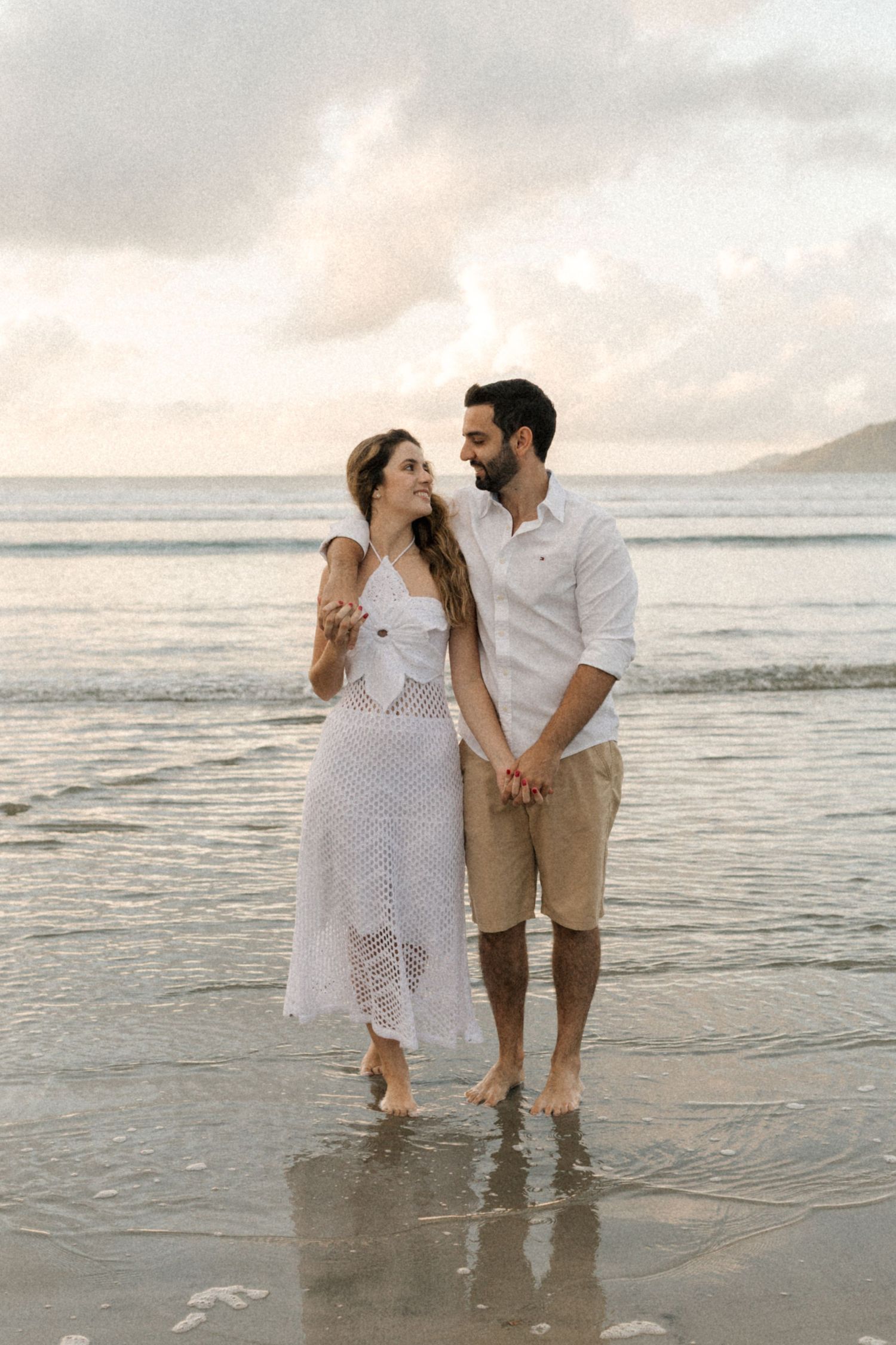 A couple walks hand in hand through shallow ocean water during sunset.