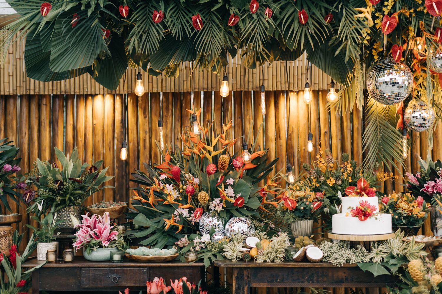 Tropical wedding dessert table with bamboo backdrop, disco balls, and lush greenery decorations.