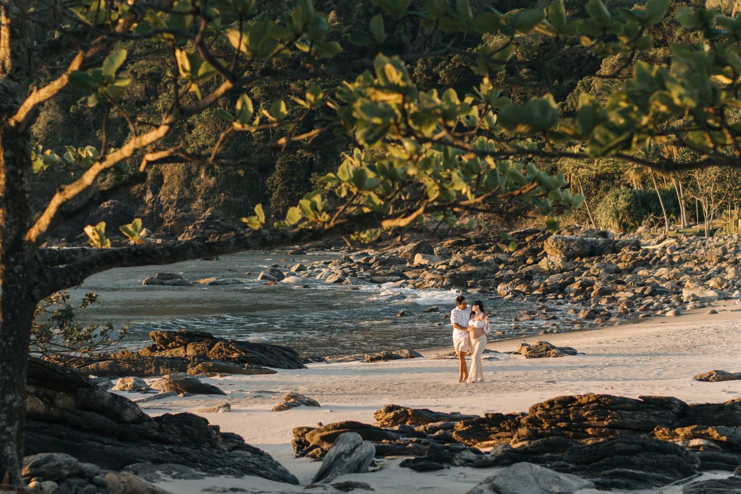 Couple strolling on sandy beach path surrounded by lush tropical foliage at sunset.