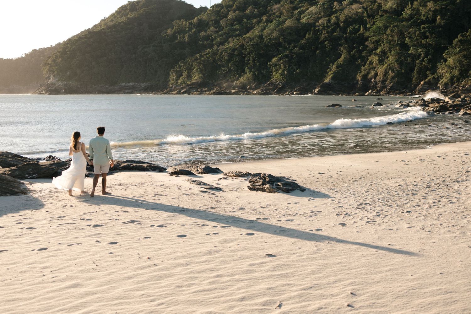 Sandy beach landscape with gentle waves rolling onto shore beneath lush green mountains in the distance.