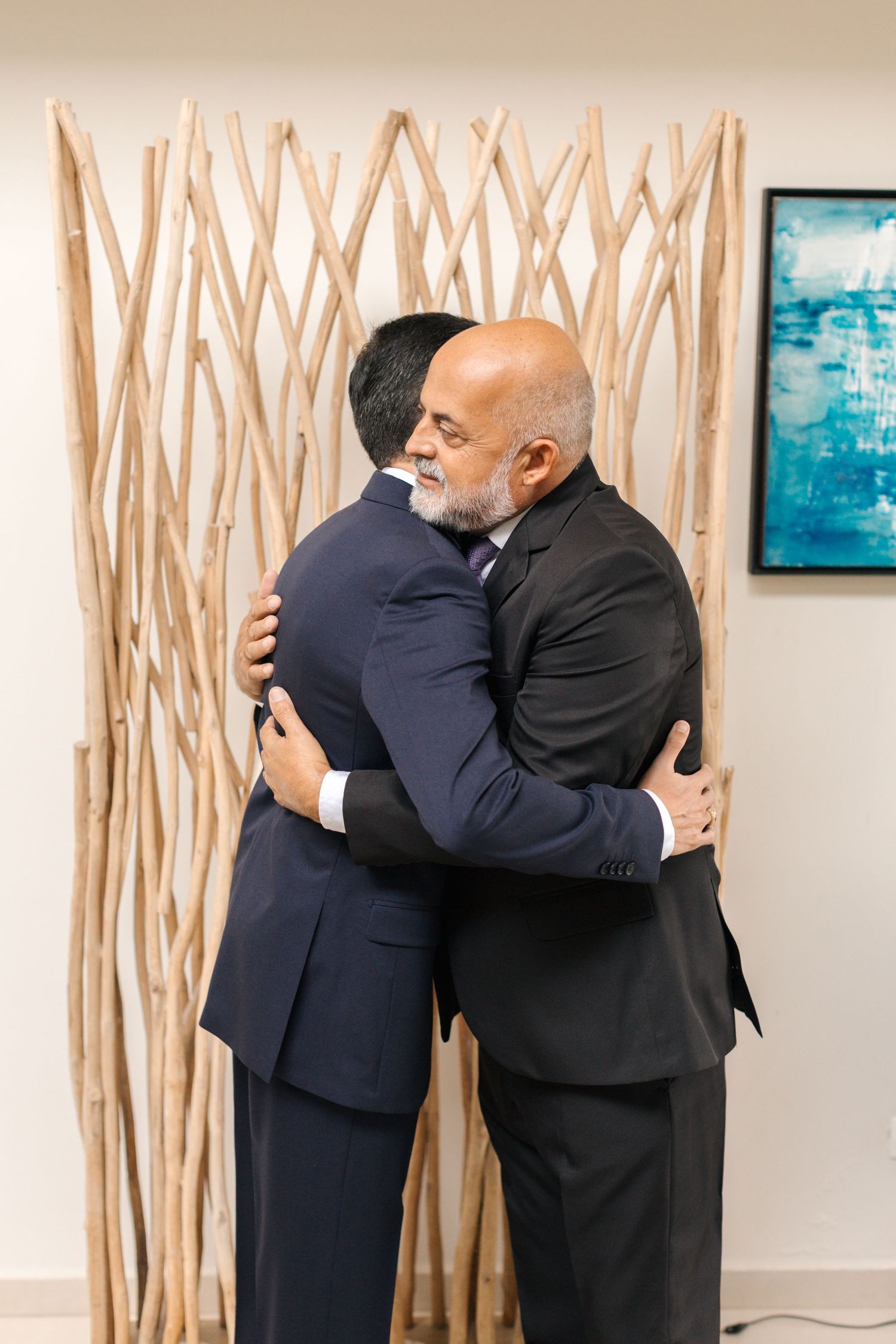 Two men in dark suits share an emotional embrace in front of a decorative driftwood wall display and blue artwork.