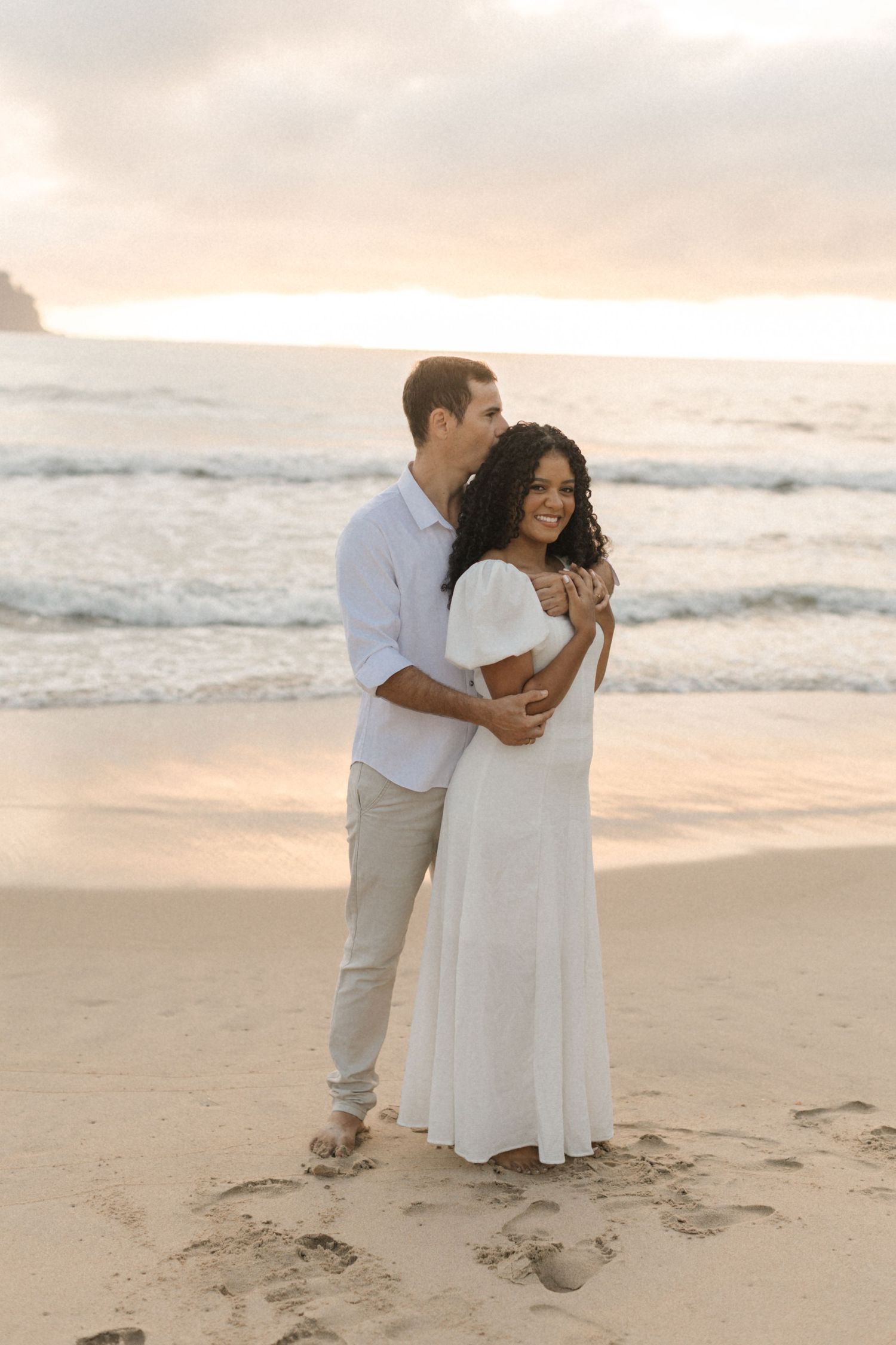 A couple embraces on a sandy beach at sunset, wearing white and casual attire against ocean waves and soft lighting.