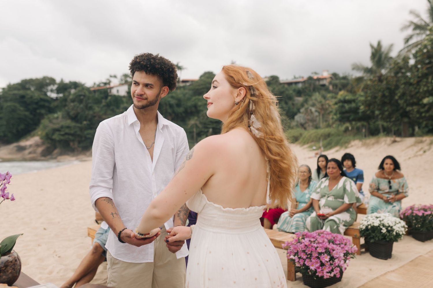 A beach ceremony scene with decorative flowers and people gathered on the sand.