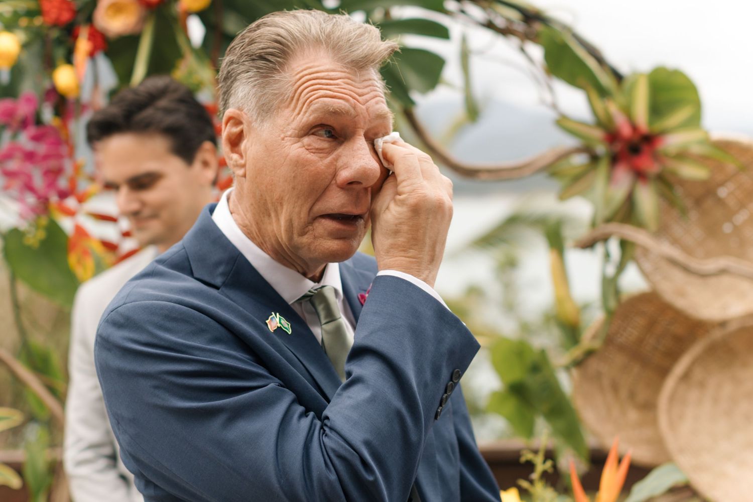 A person in a navy suit stands among tropical floral arrangements at an outdoor venue.