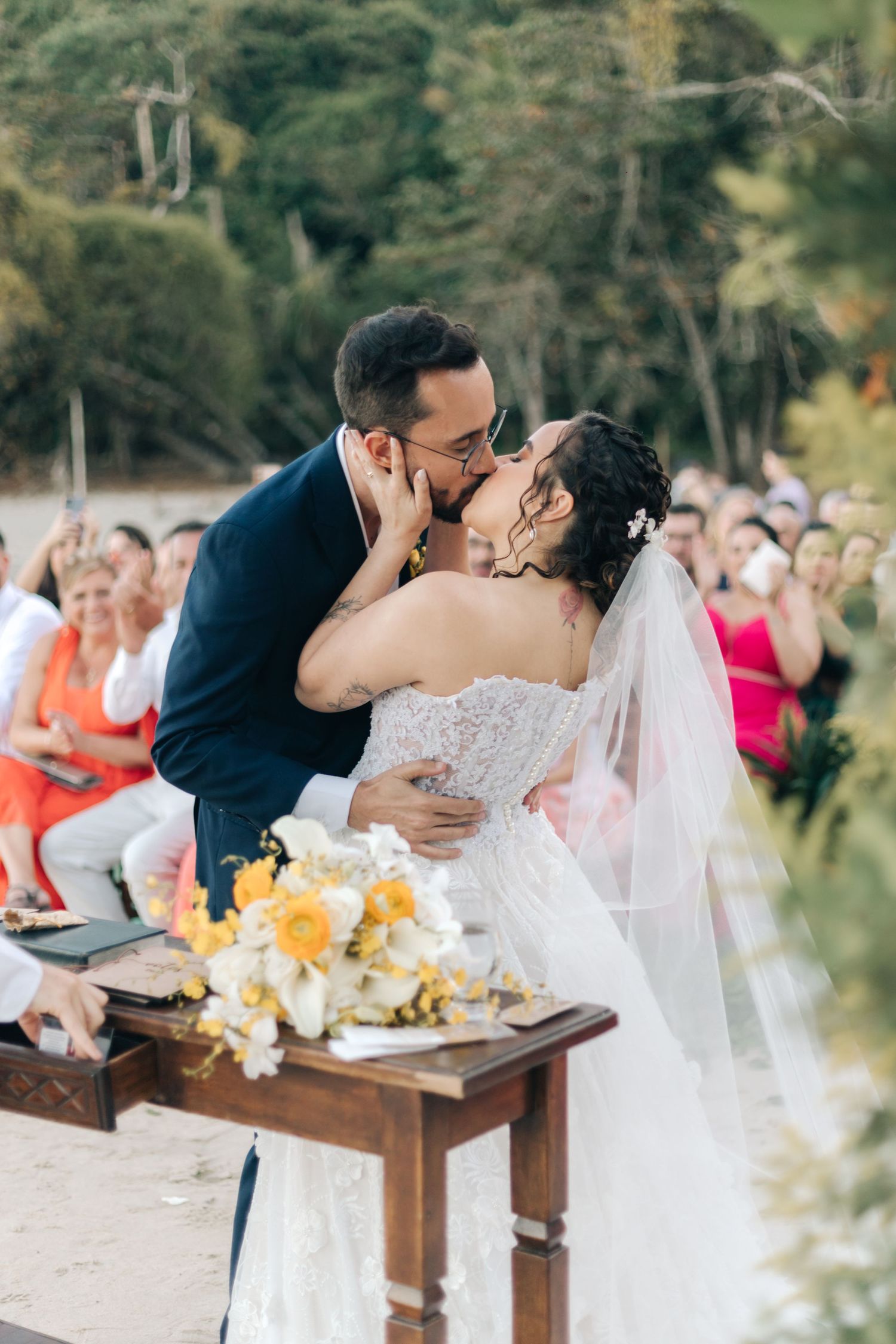 Couple shares romantic kiss at beach wedding altar with guests watching.