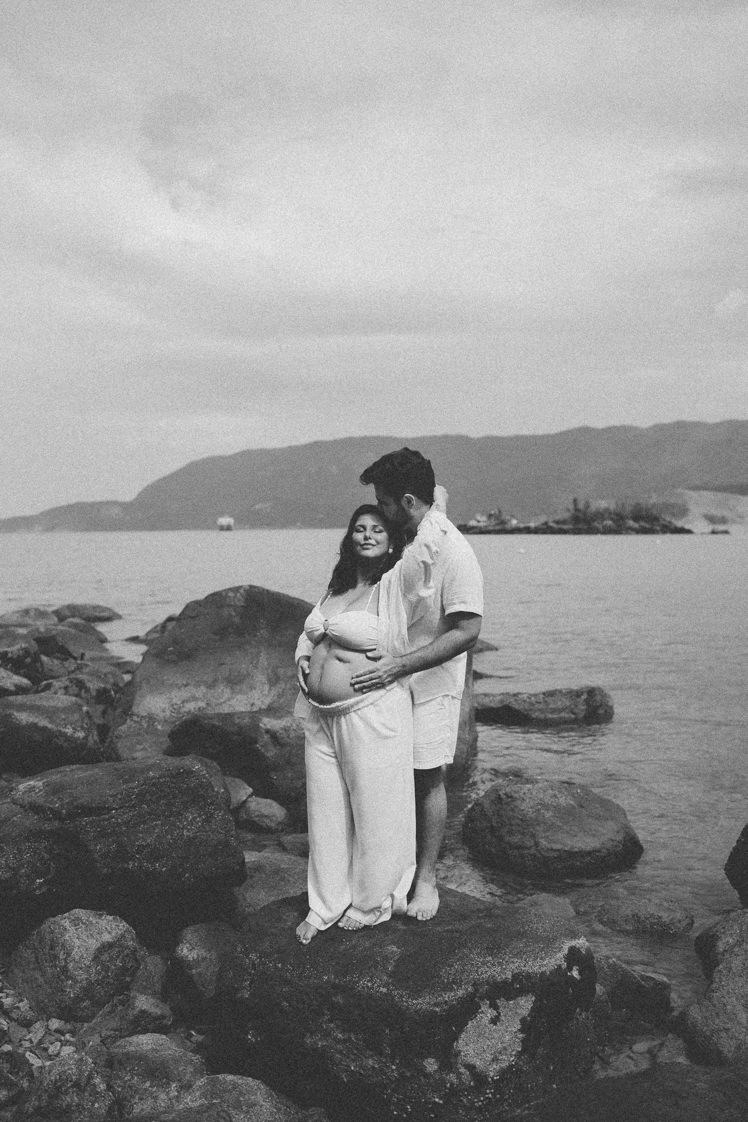 A couple stands on rocky coastal cliffs overlooking the ocean in black and white photography.