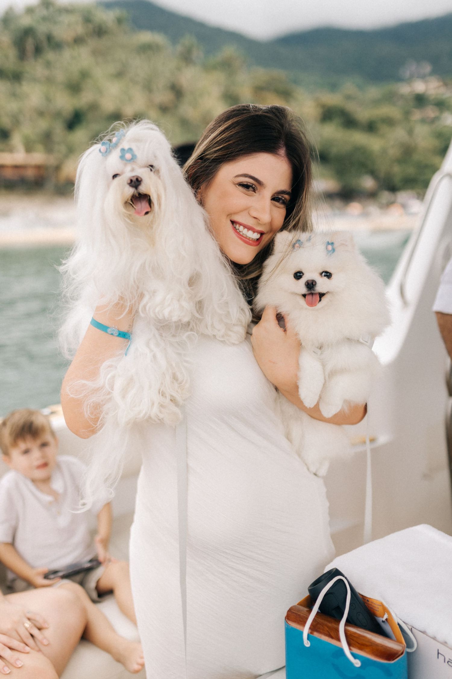 Person holding two fluffy white dogs on boat with mountain scenery in background.