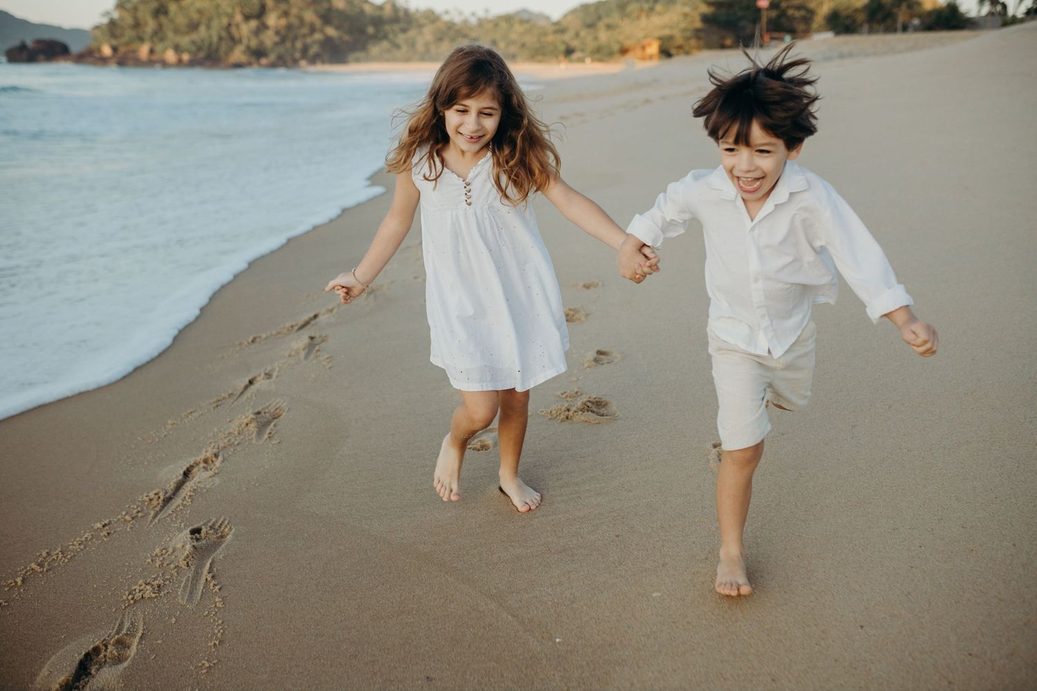 People in light clothing skip along the seashore with mountains visible in the background.