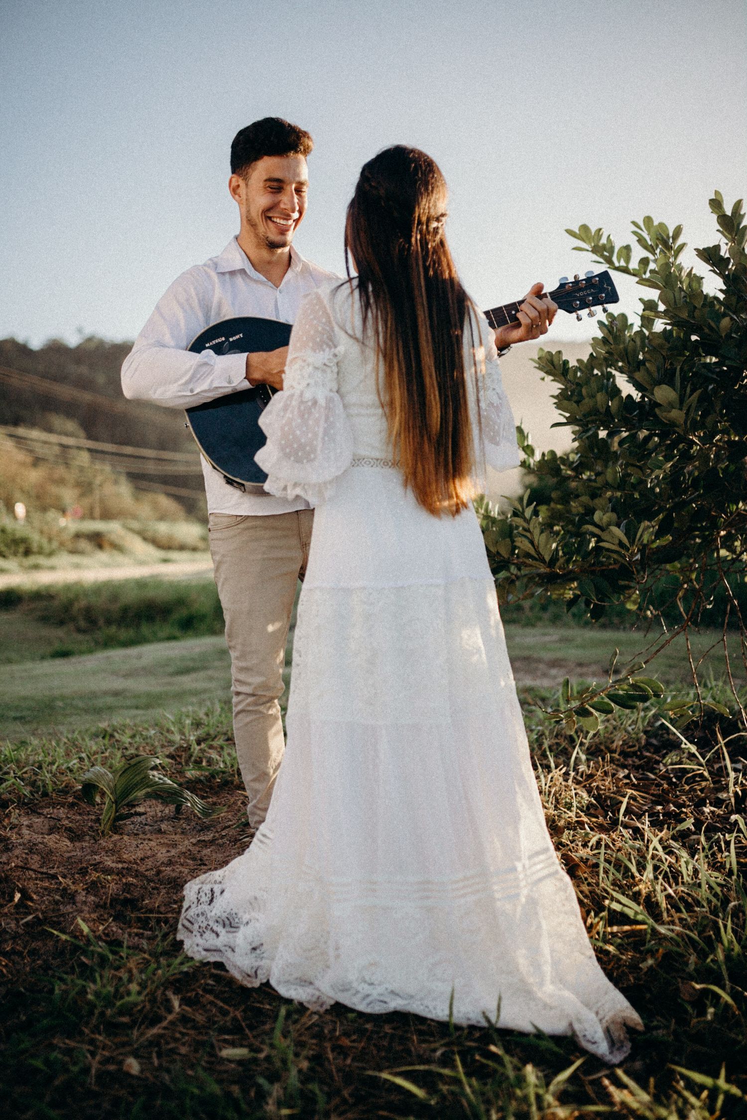 Couple in wedding attire pose outdoors with a guitar amidst scenic hills and greenery.