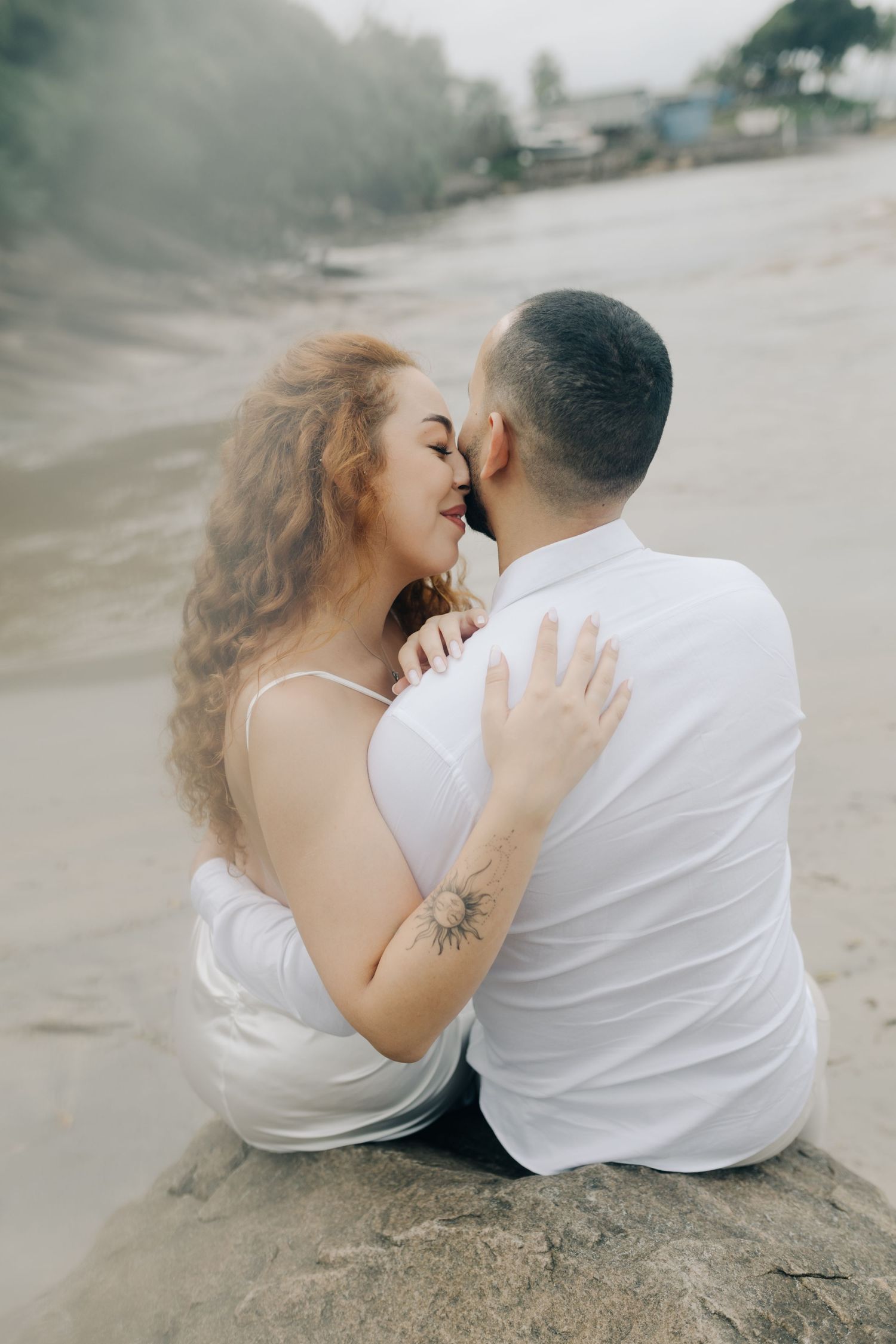 Couple in white sitting on beach sand sharing intimate moment.