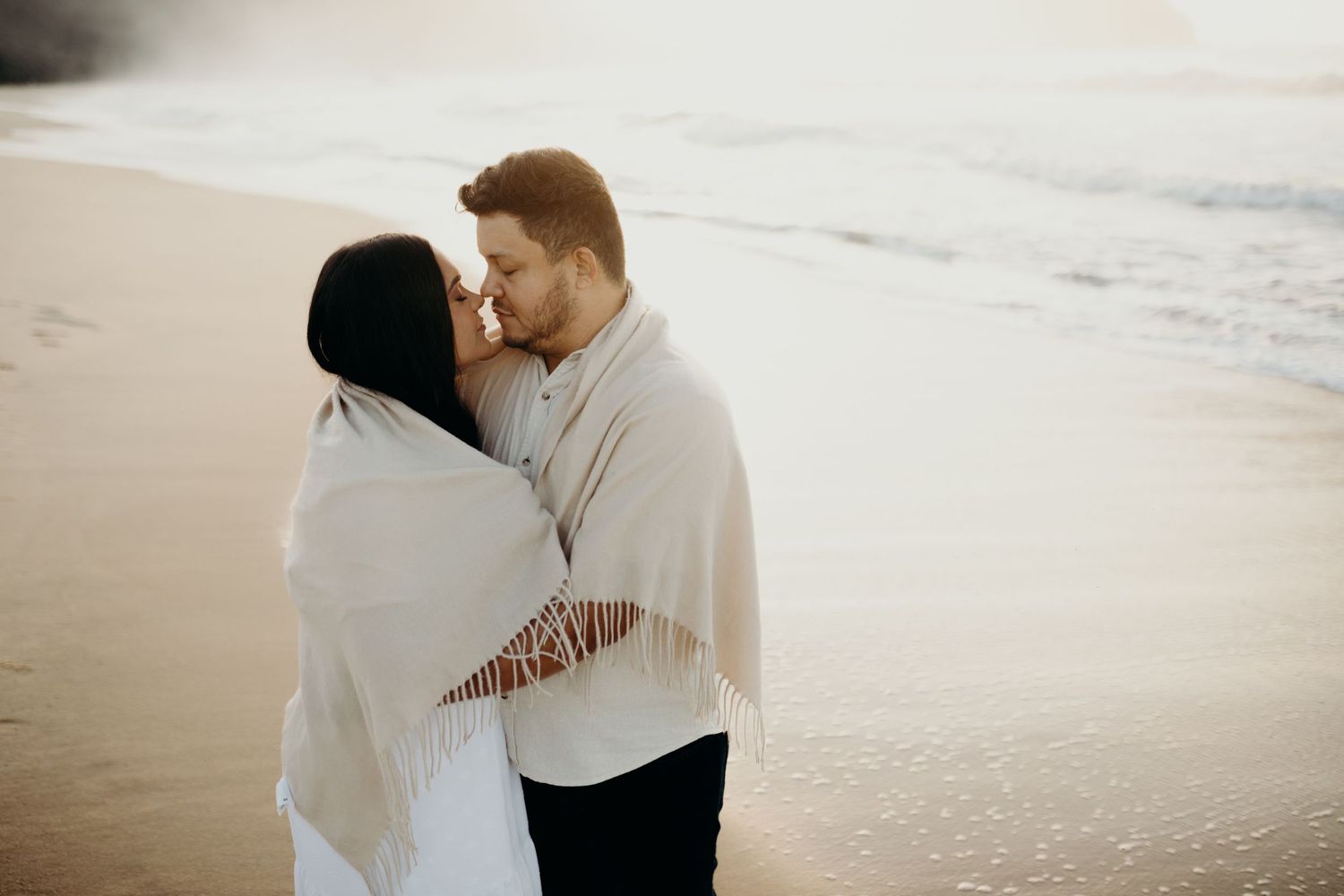 Couple embraces while wrapped in a fringed white shawl at the beach.