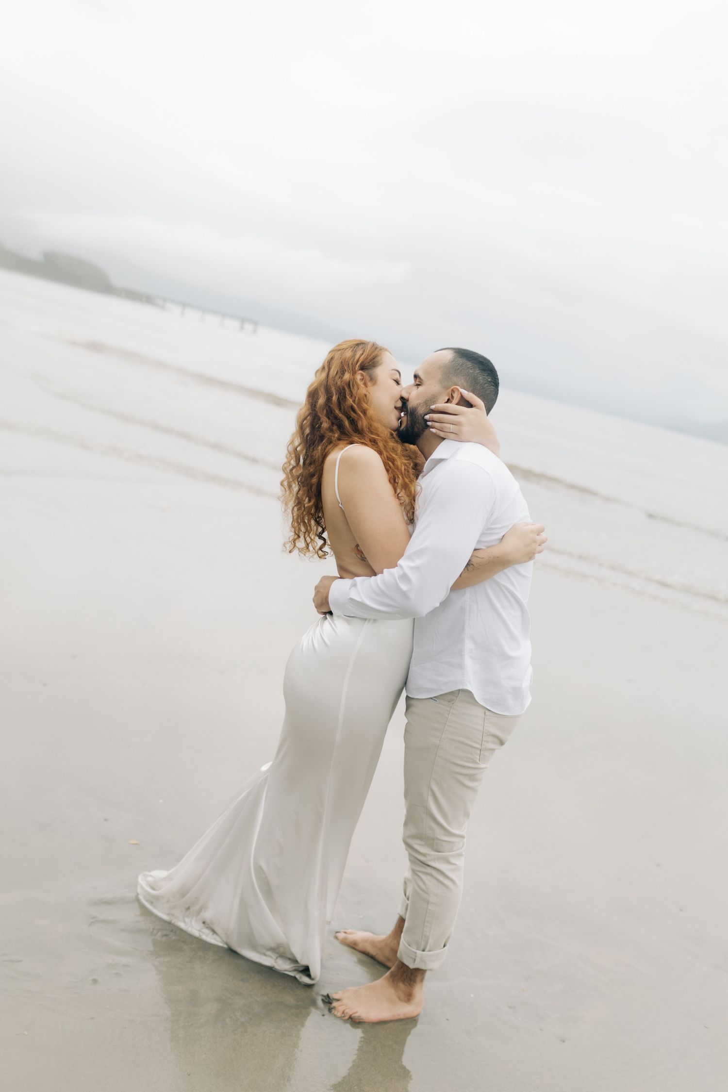 Couple in white shares romantic kiss on foggy beach during engagement shoot.