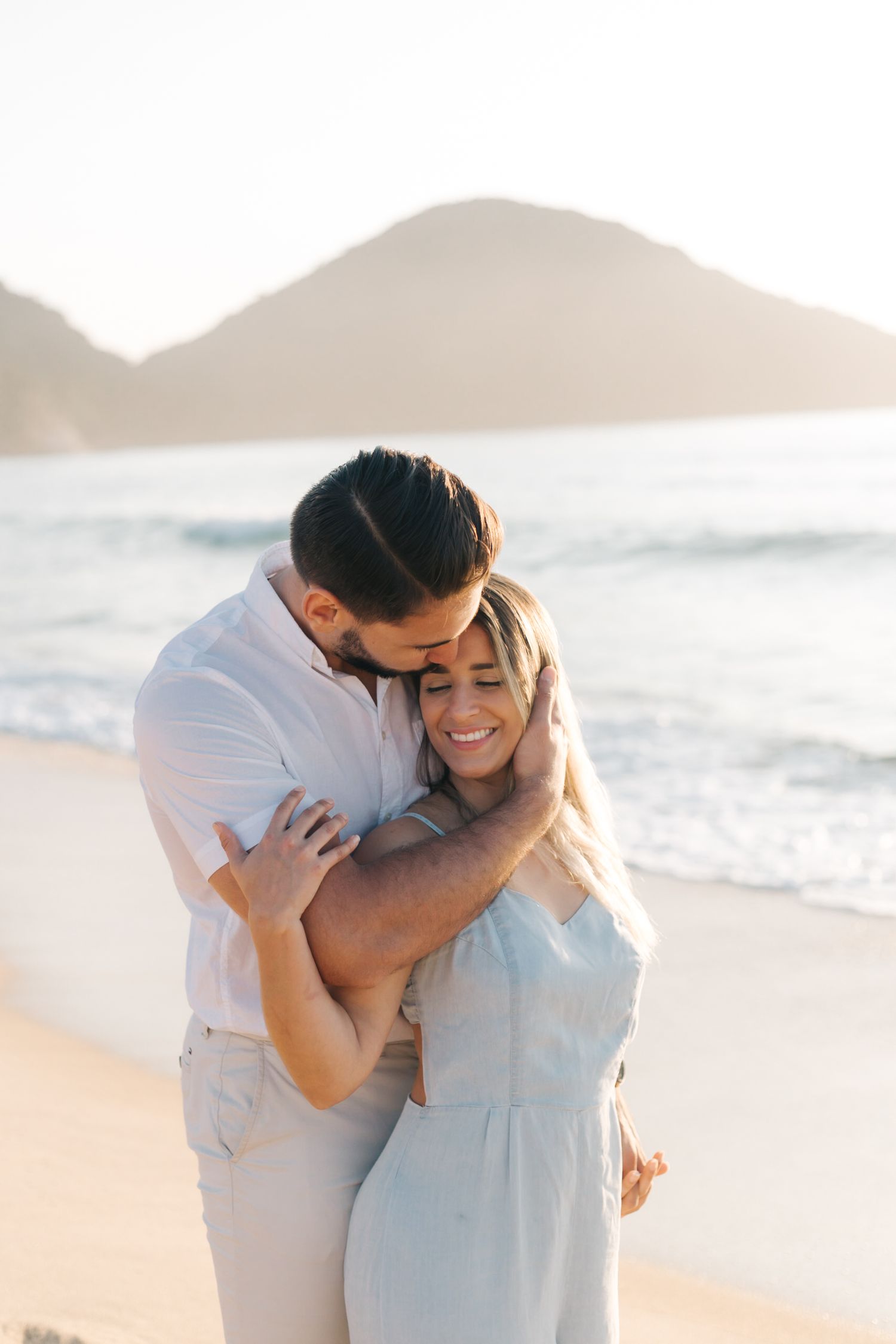Intimate moment between couple hugging on sandy beach with coastal mountains and waves.