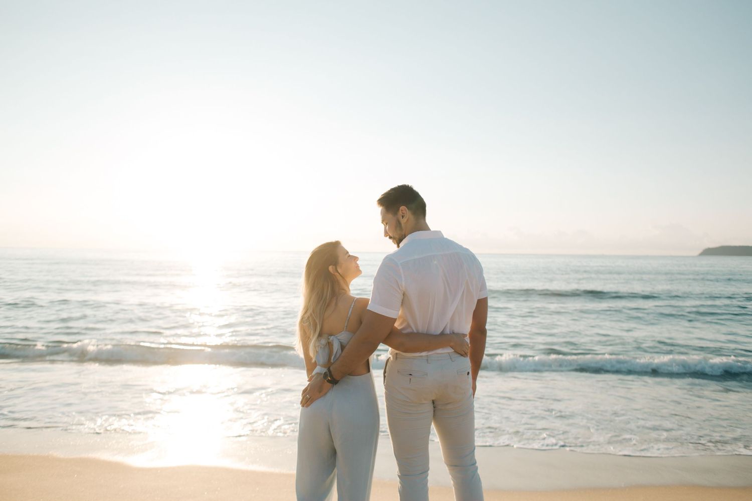 Two people in white outfits sharing intimate moment on beach at sunset.