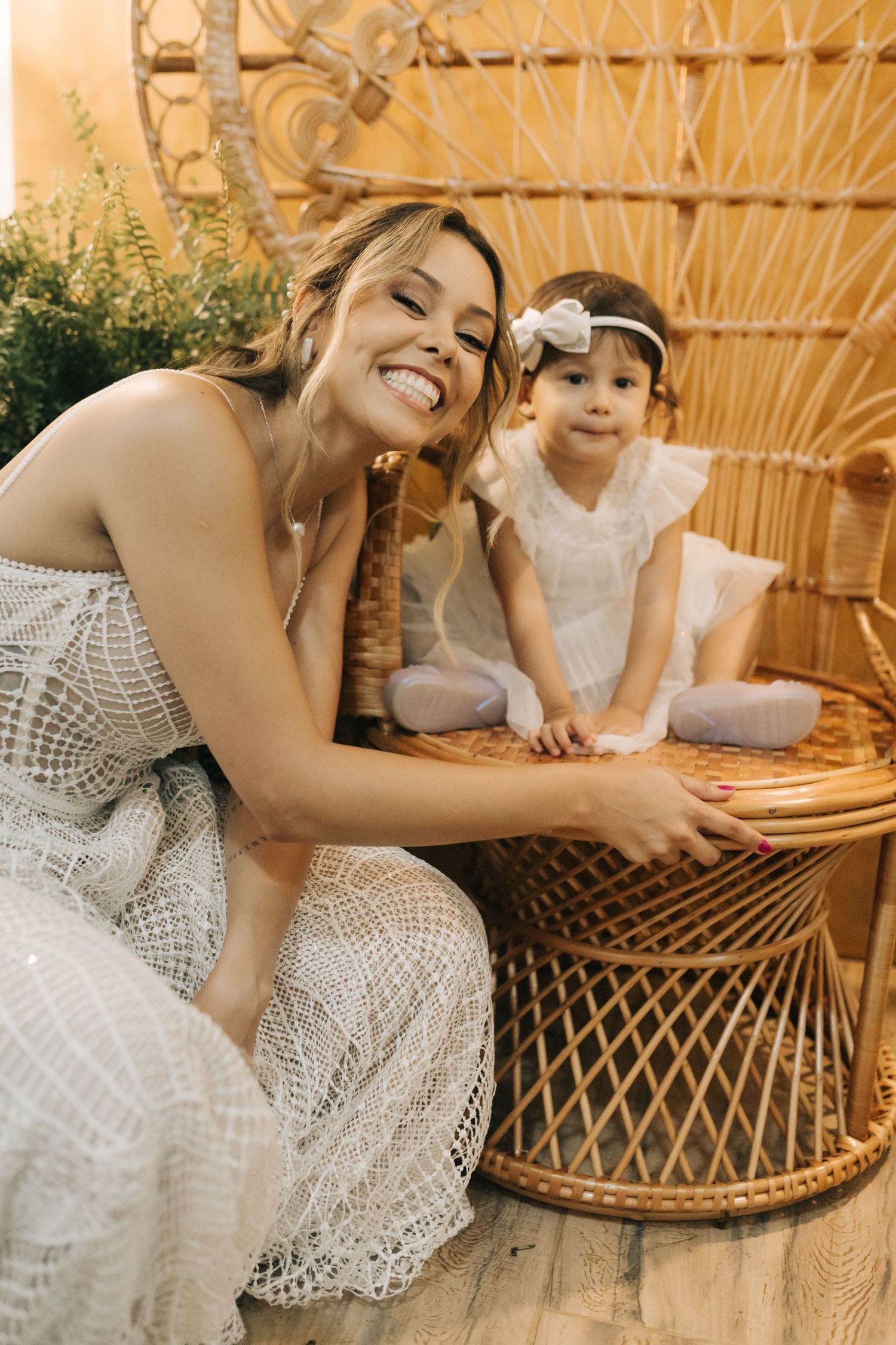 Bride in lace dress sharing sweet moment with flower girl on woven peacock chair.