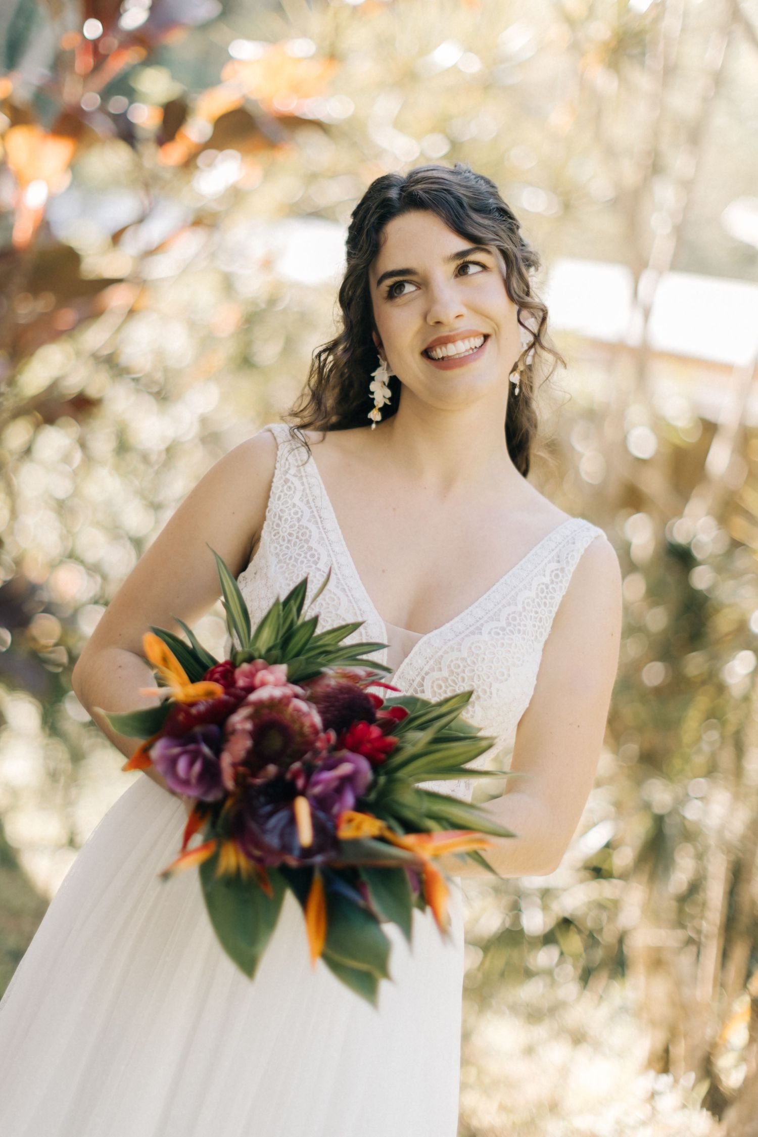Bride in a white v-neck dress holds a vibrant tropical bouquet in a sun-dappled garden setting.