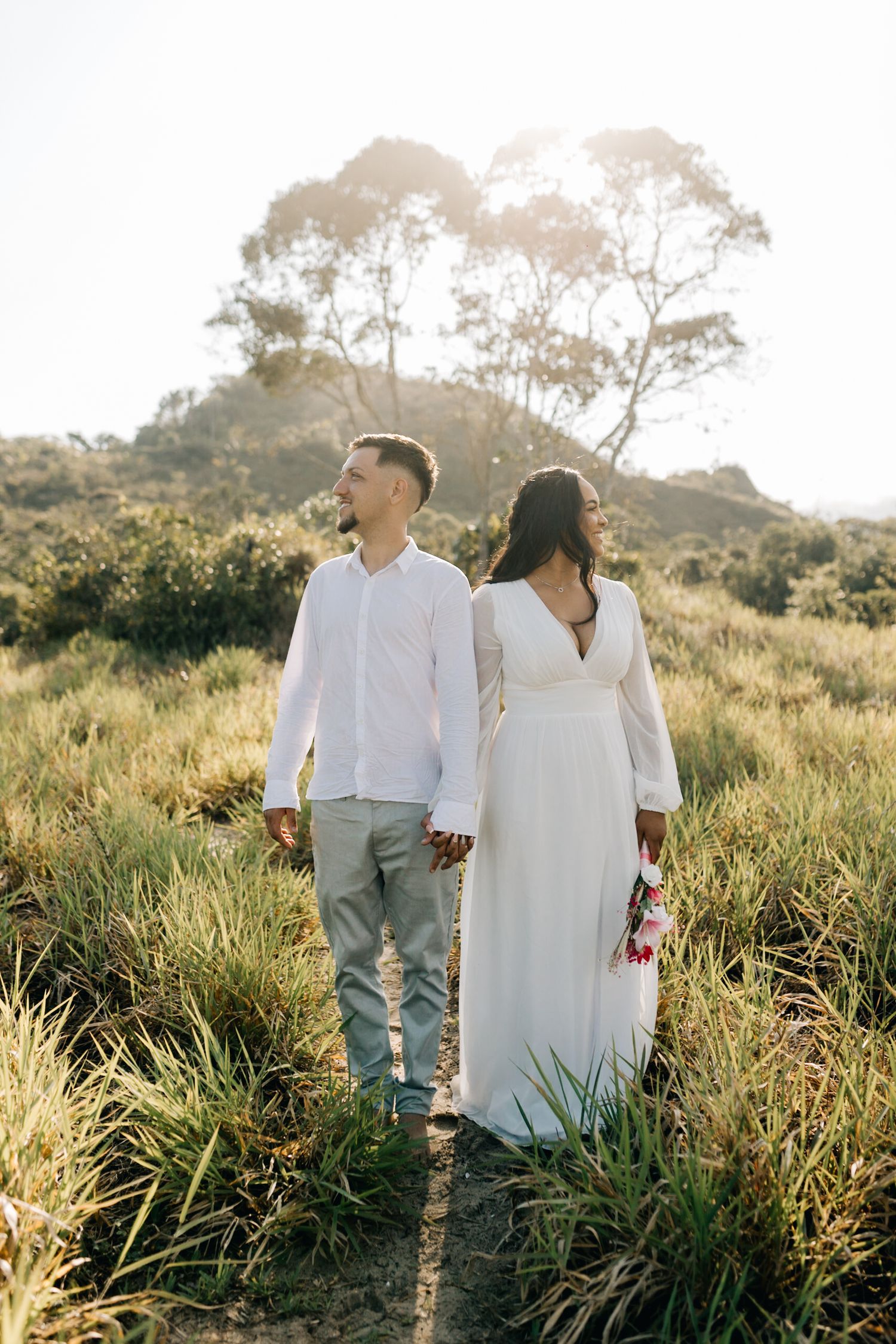 Romantic couple strolling through a sun-lit meadow wearing coordinated white outfits.