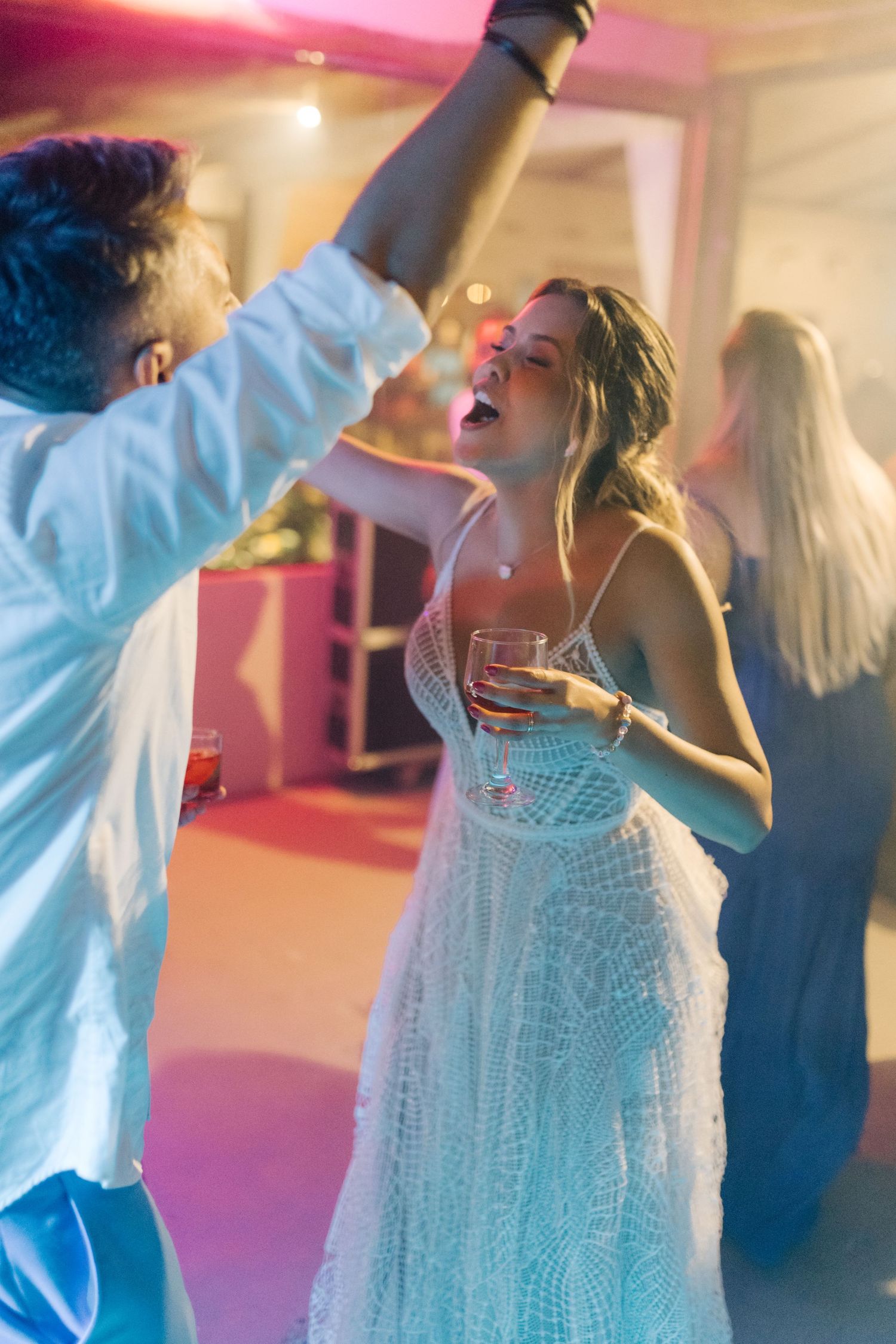 Couple dancing energetically at a wedding reception with colorful lighting and the bride in a white lace dress.