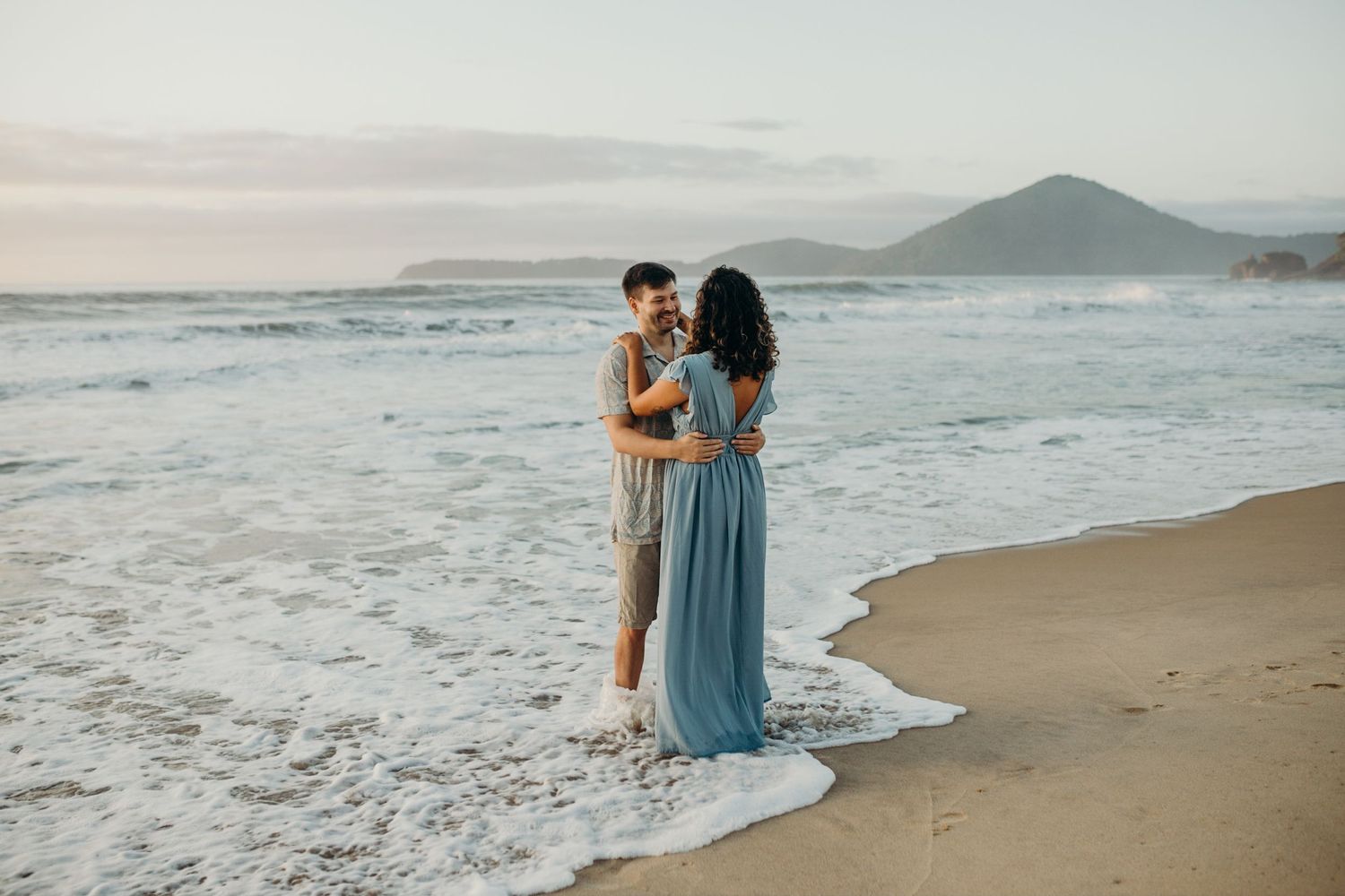 Couple walking along scenic coastal beach with mountains and waves in background.