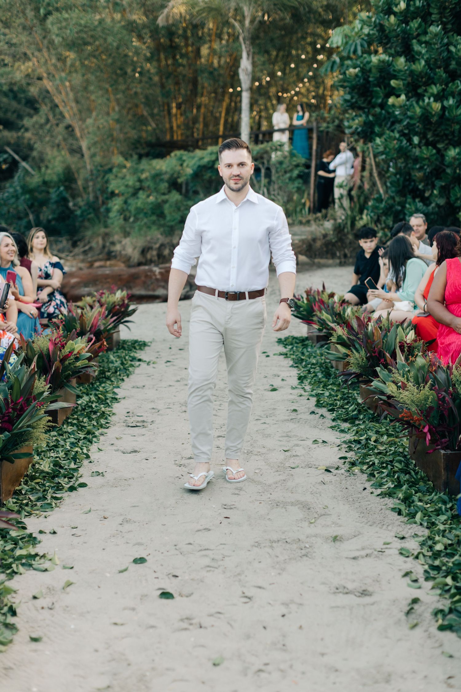 Groomsman in white shirt and beige pants walks sandy aisle at garden wedding.