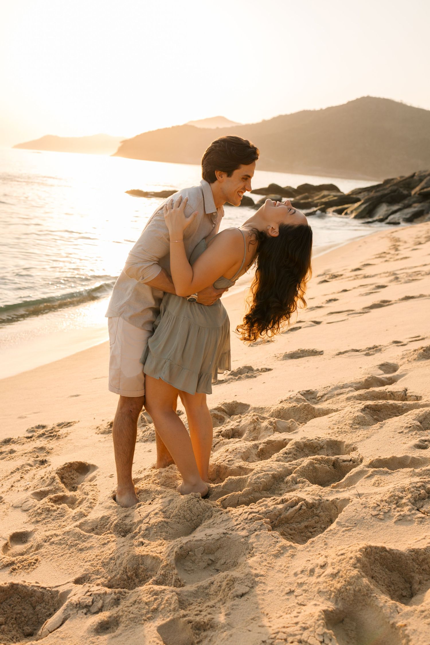Playful couple embracing and laughing on a sandy beach at sunset with mountains in the background.