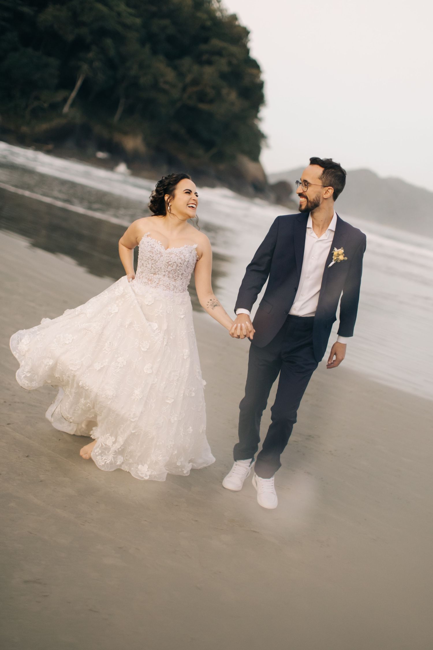 A couple walks hand in hand on a beach, with the bride wearing a flowing lace gown and groom in a navy suit.