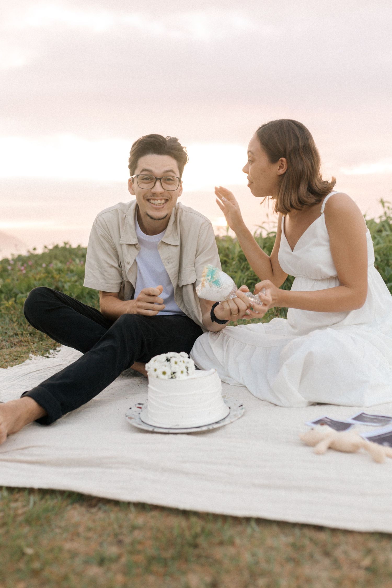 A couple shares an intimate picnic with cake on a blanket during sunset in a grassy field.