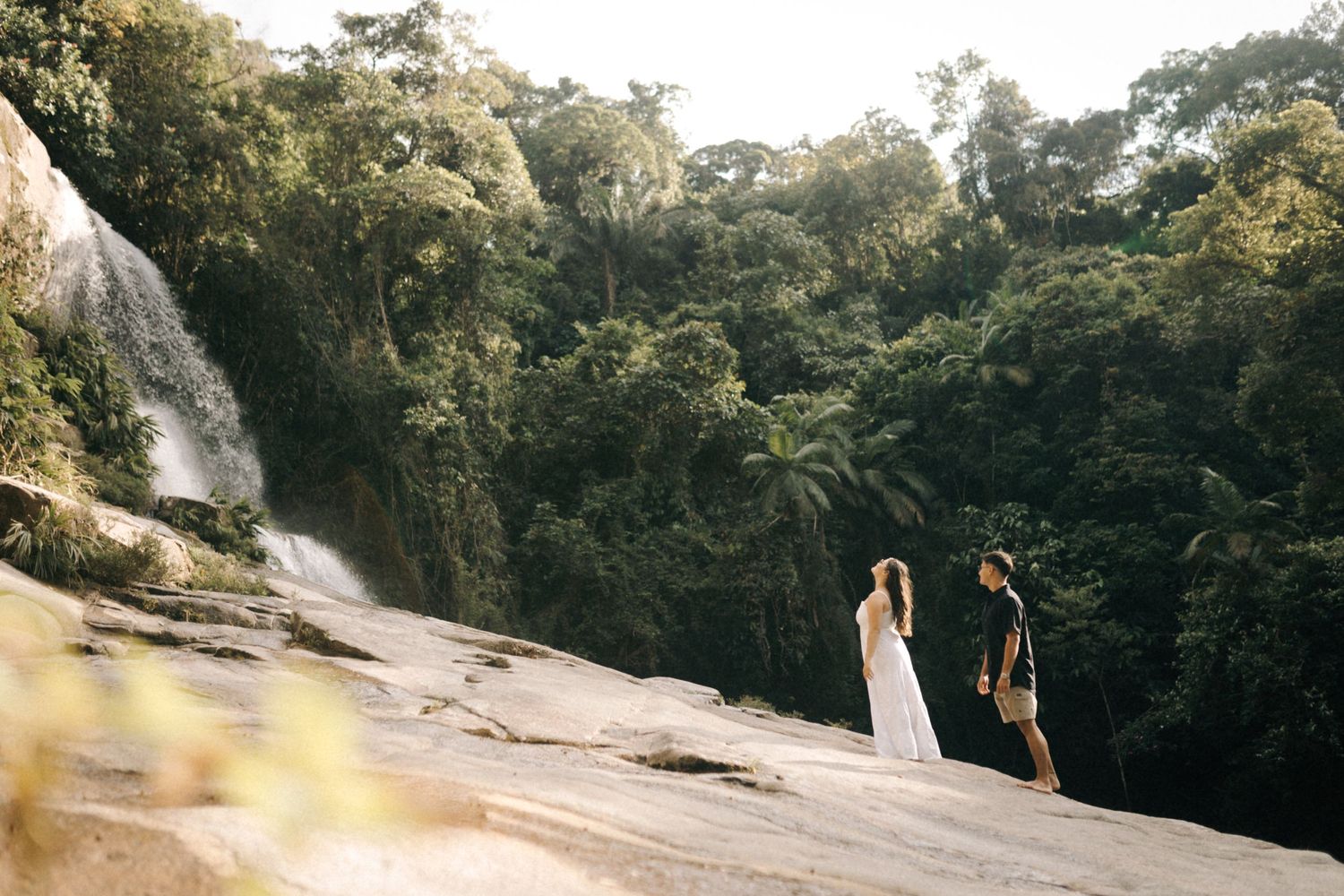 um casal estão em uma encosta rochosa perto de uma cachoeira cercada por uma exuberante floresta verde.