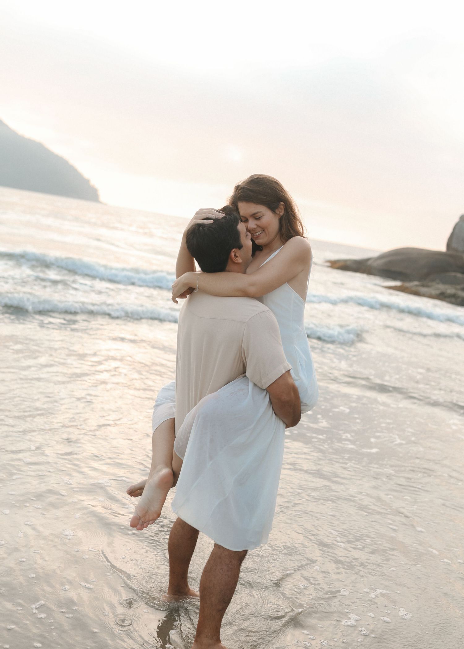 A tender beach photoshoot showing a joyful lift pose with ocean waves in the background.