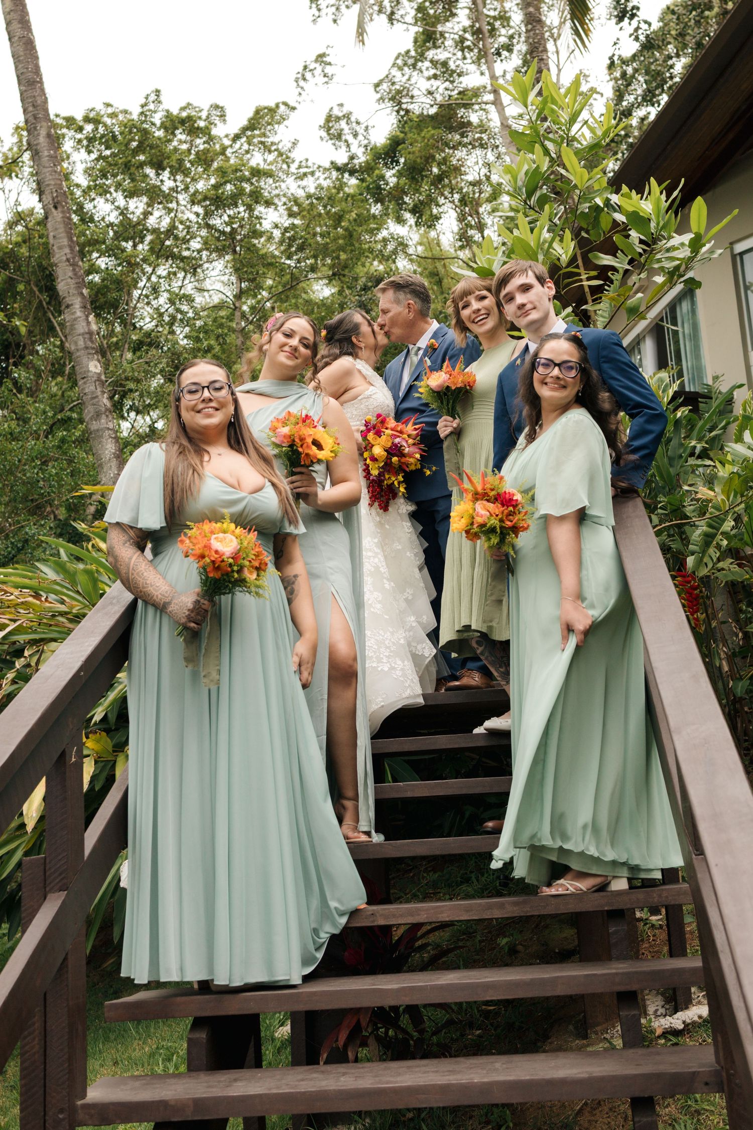 Wedding party in mint green dresses poses on wooden stairs surrounded by tropical foliage.