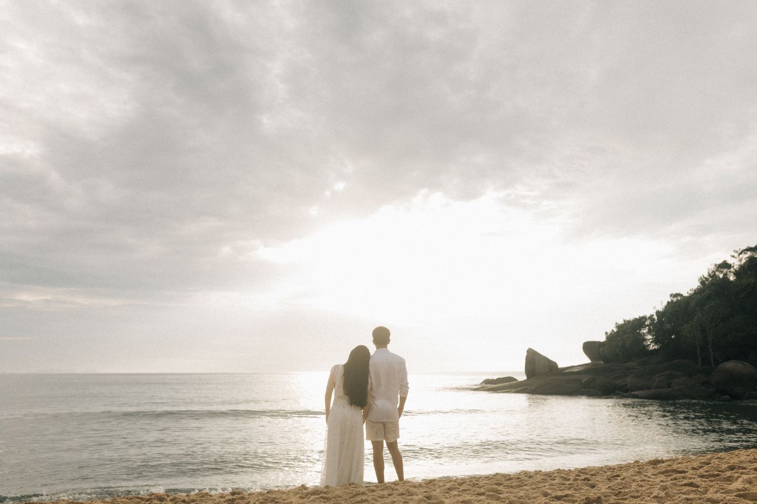 couple stands silhouetted against a sunset on a peaceful beach with rocky cliffs in the background.