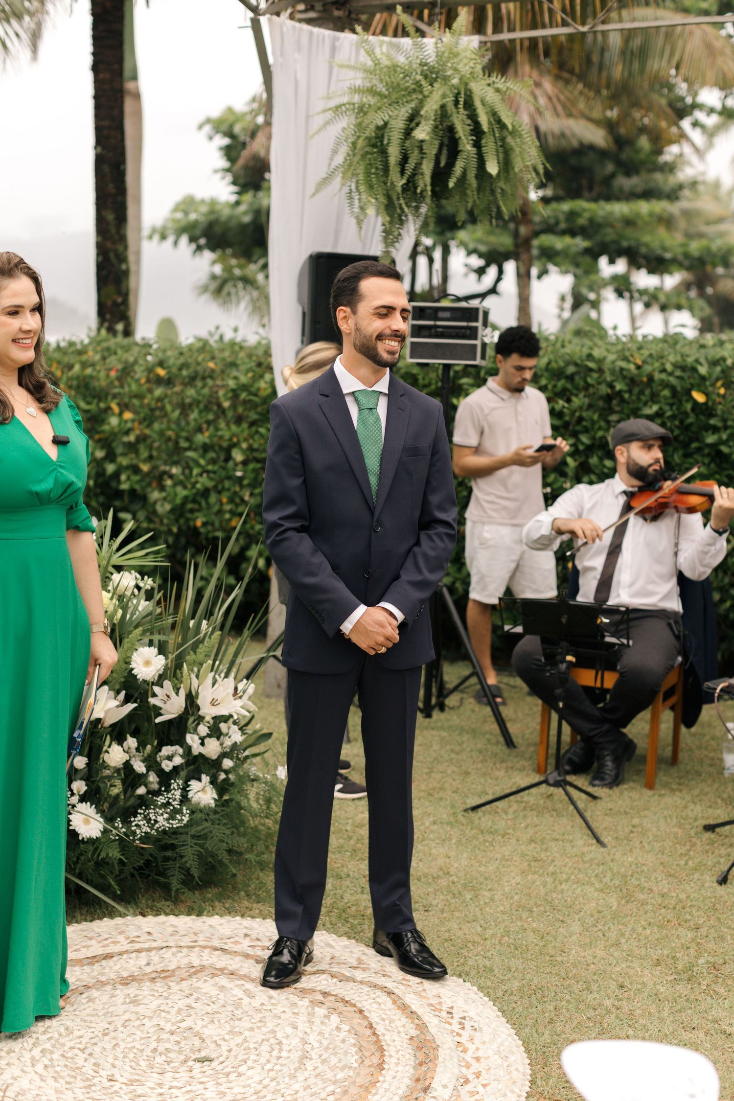 Wedding party members in navy suits and emerald green dresses stand on a decorated outdoor pathway.