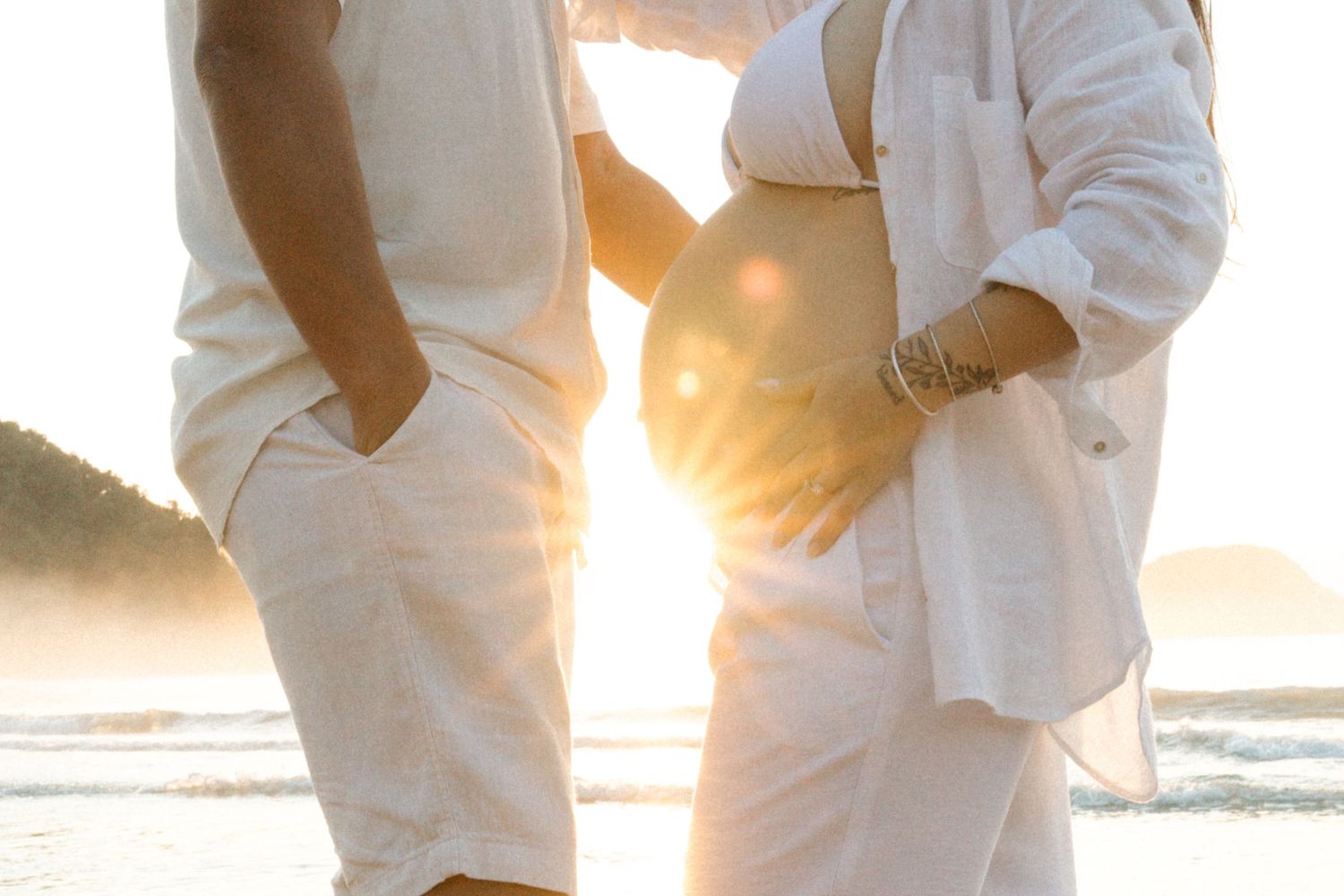 Expectant couple in white clothing silhouetted against golden sunset on beach.