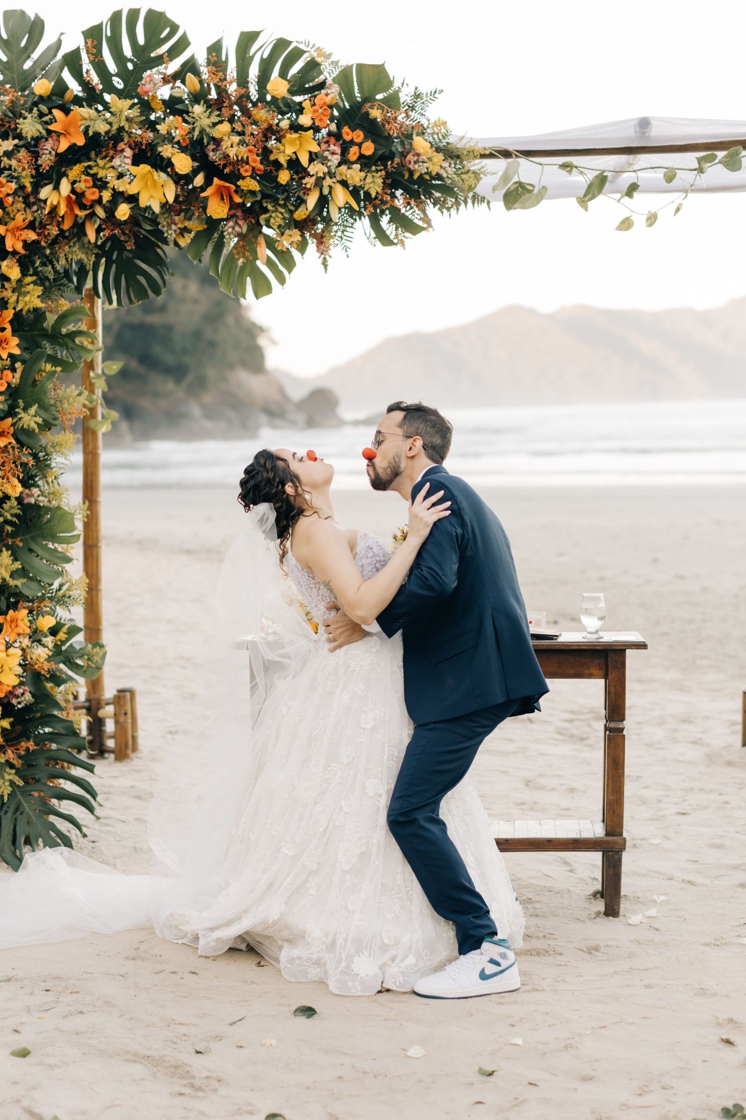 Dancing newlyweds share a romantic moment beneath an orange and green floral arch on the beach.