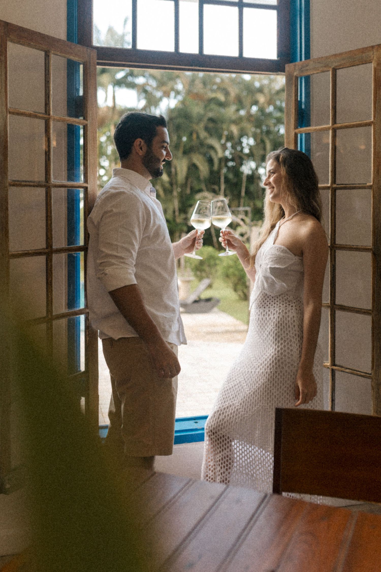 A romantic couple toasts with wine glasses while standing in an open doorway with natural light streaming in.