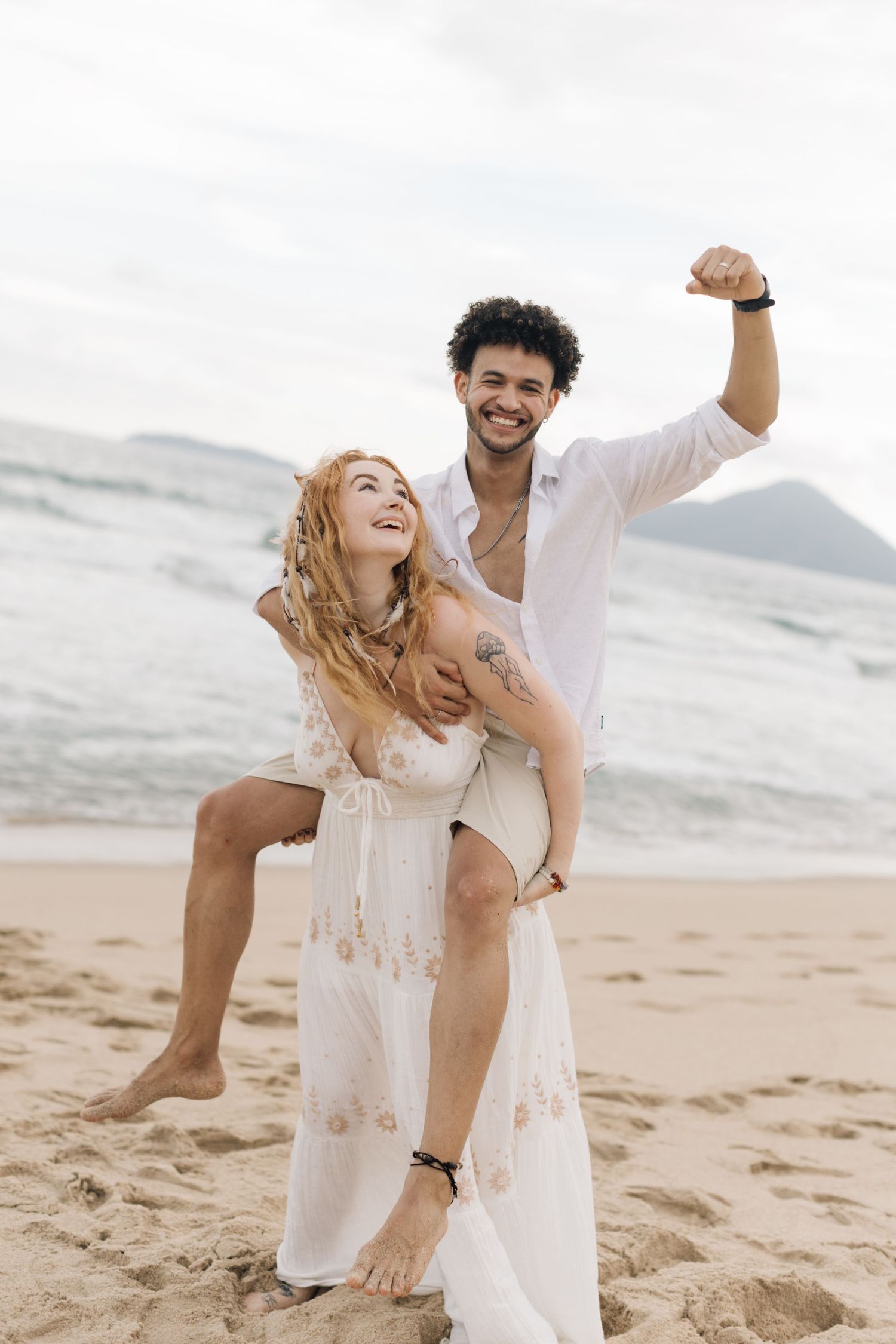 A playful couple in white clothing dances and laughs together on a sandy beach.