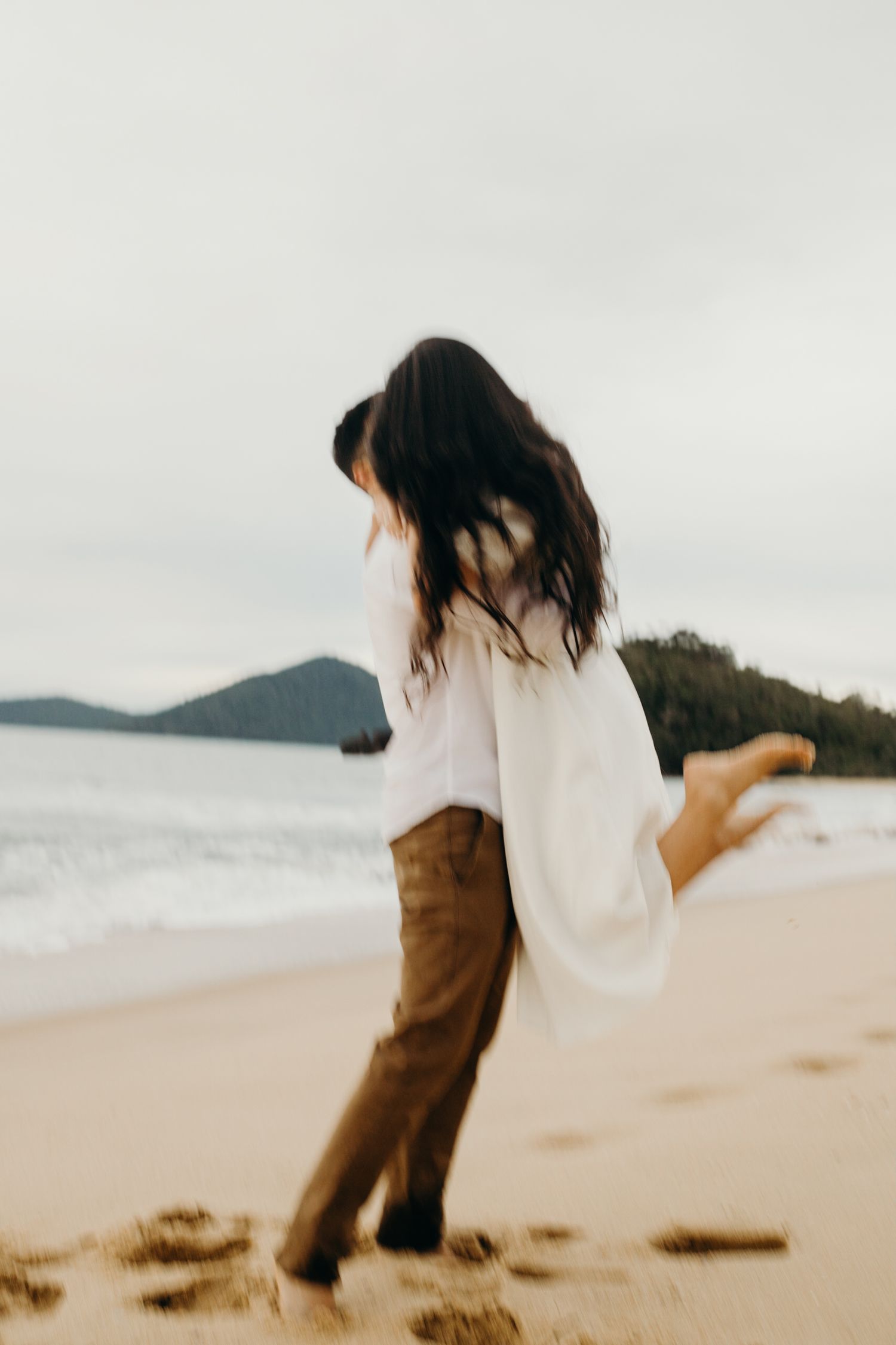 A couple shares a joyful moment on a scenic beach with mountains in the background.