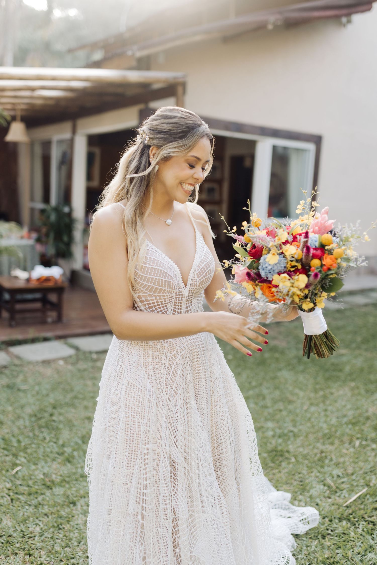 A radiant bride in a lace gown admires her colorful wedding bouquet in an outdoor garden setting.