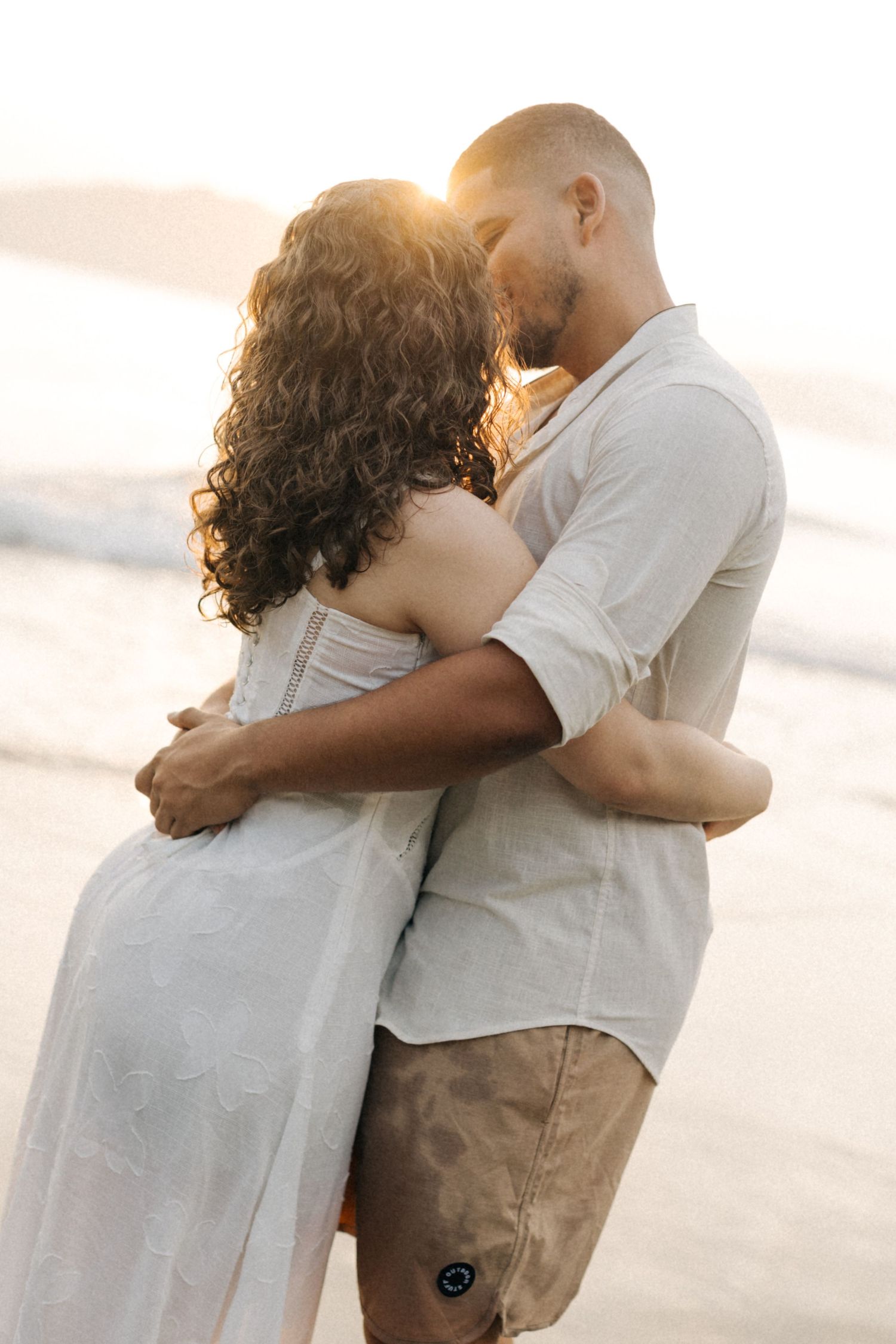A couple embraces on the beach during golden hour with sunlit waves behind them.