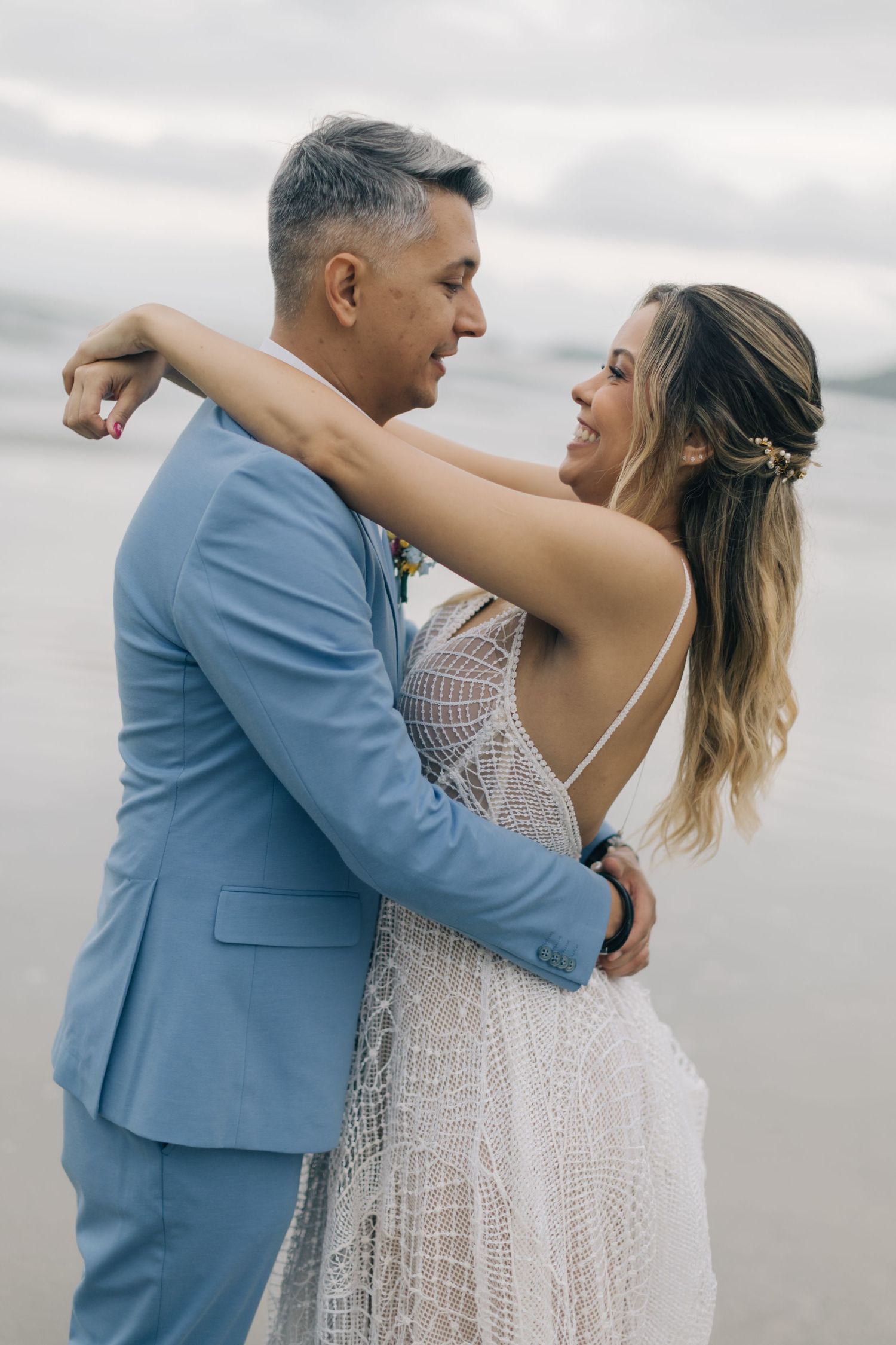 A couple in blue suit and white lace dress embrace during their beach wedding photos.
