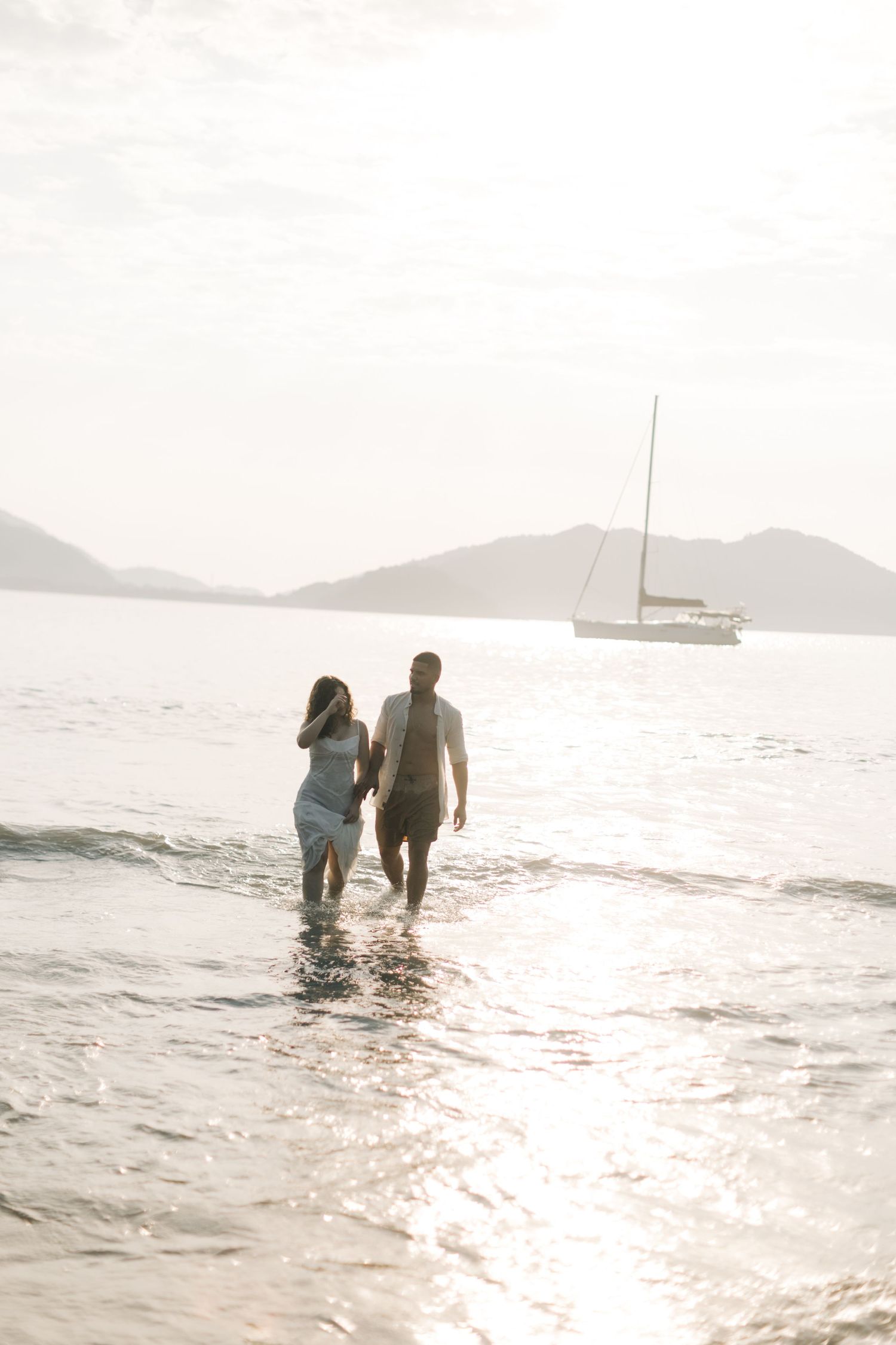 A couple walking hand in hand in shallow ocean water with a sailboat in the distance.