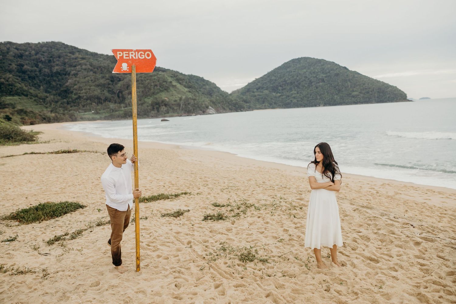 A scenic beach engagement photo with mountains and warning signs in the background.