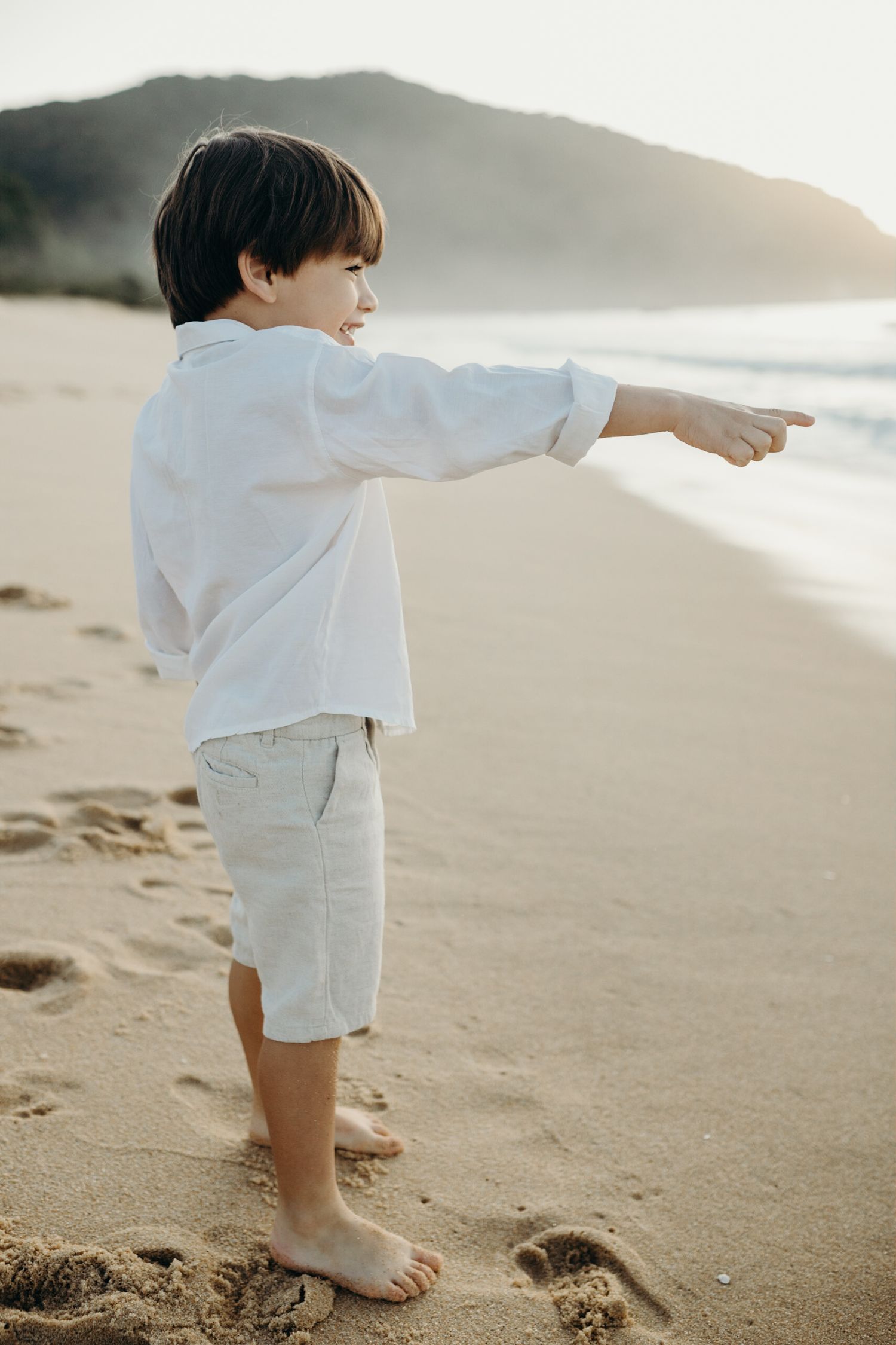 Small child wearing white outfit standing on sandy beach at sunset looking toward the ocean.