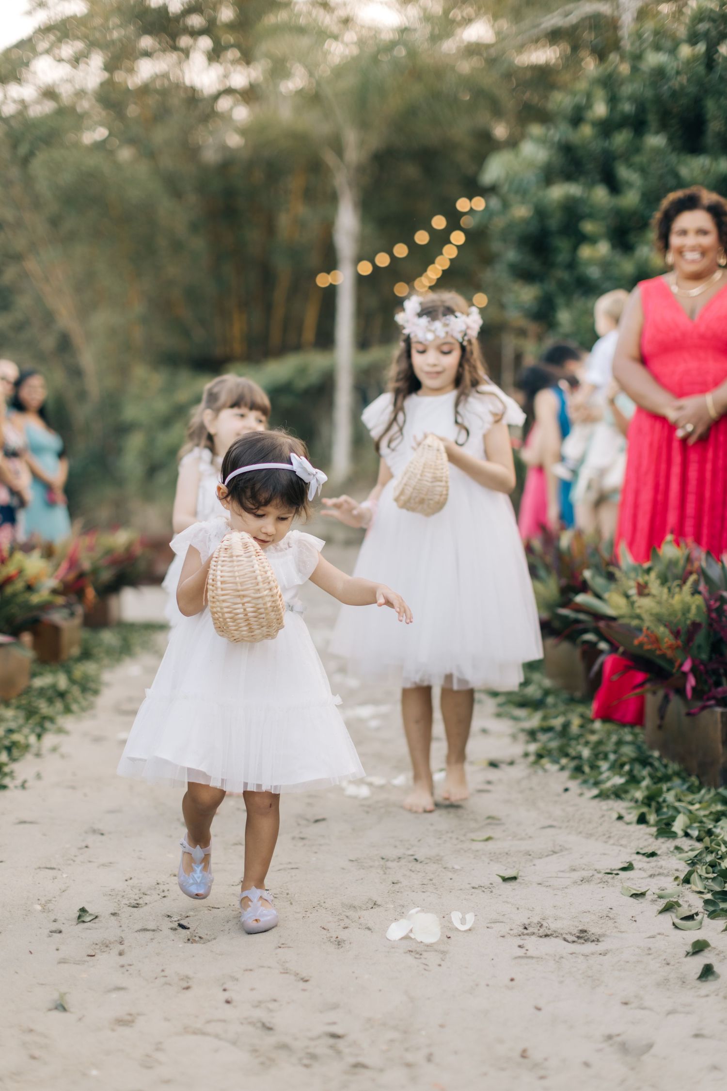 Young flowergirl in white dress playfully holding a wicker basket while walking down a garden pathway.