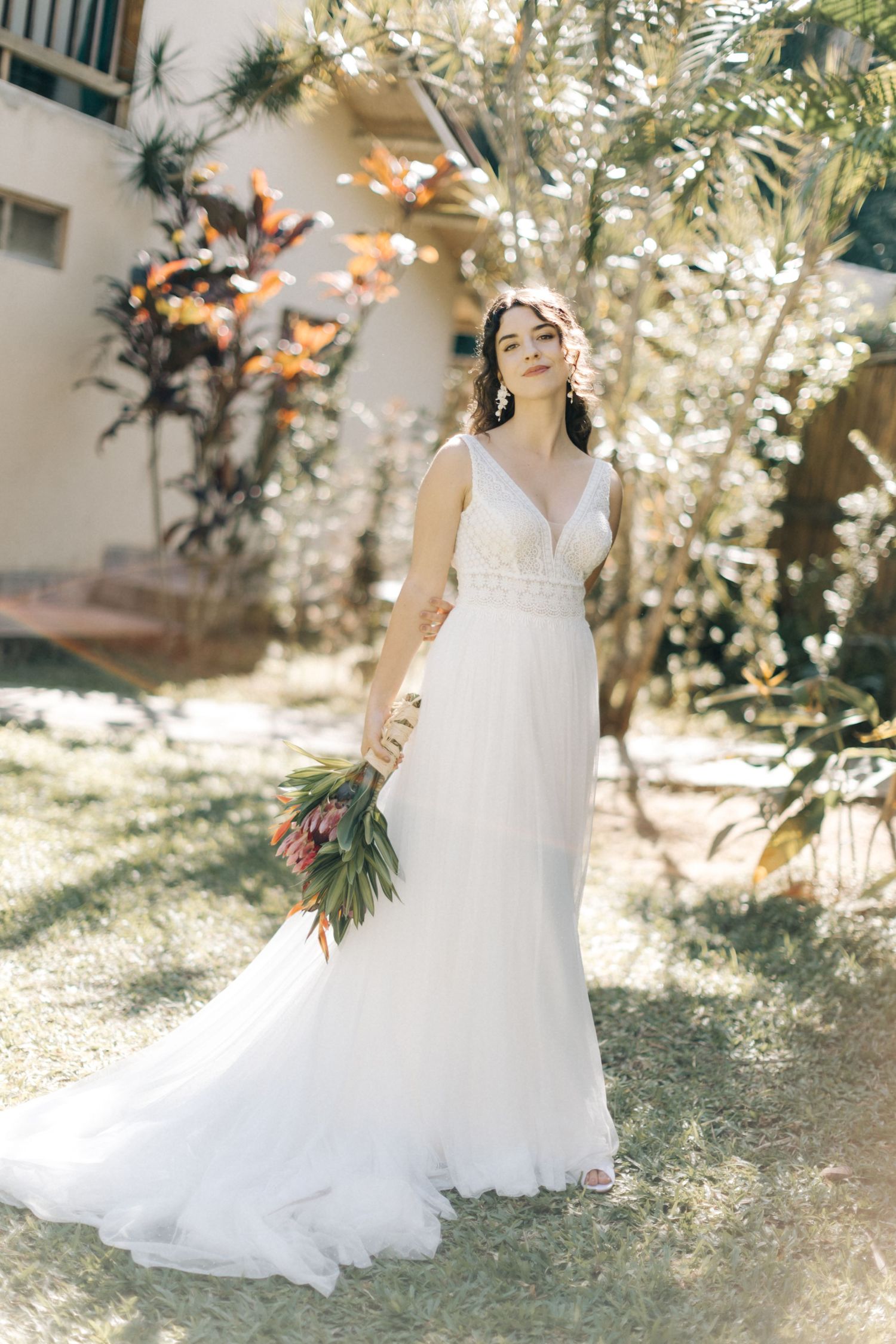 A bride in an elegant white gown standing in dappled sunlight with tropical flowers.