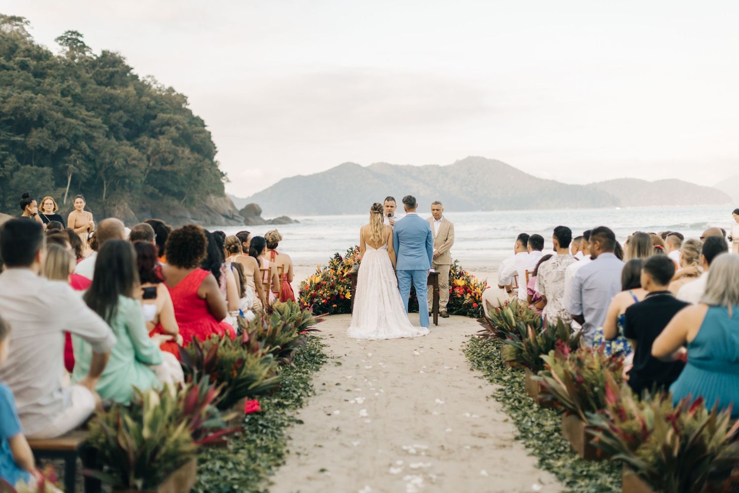 Wedding guests gather at an outdoor coastal venue with mountain views in the background.