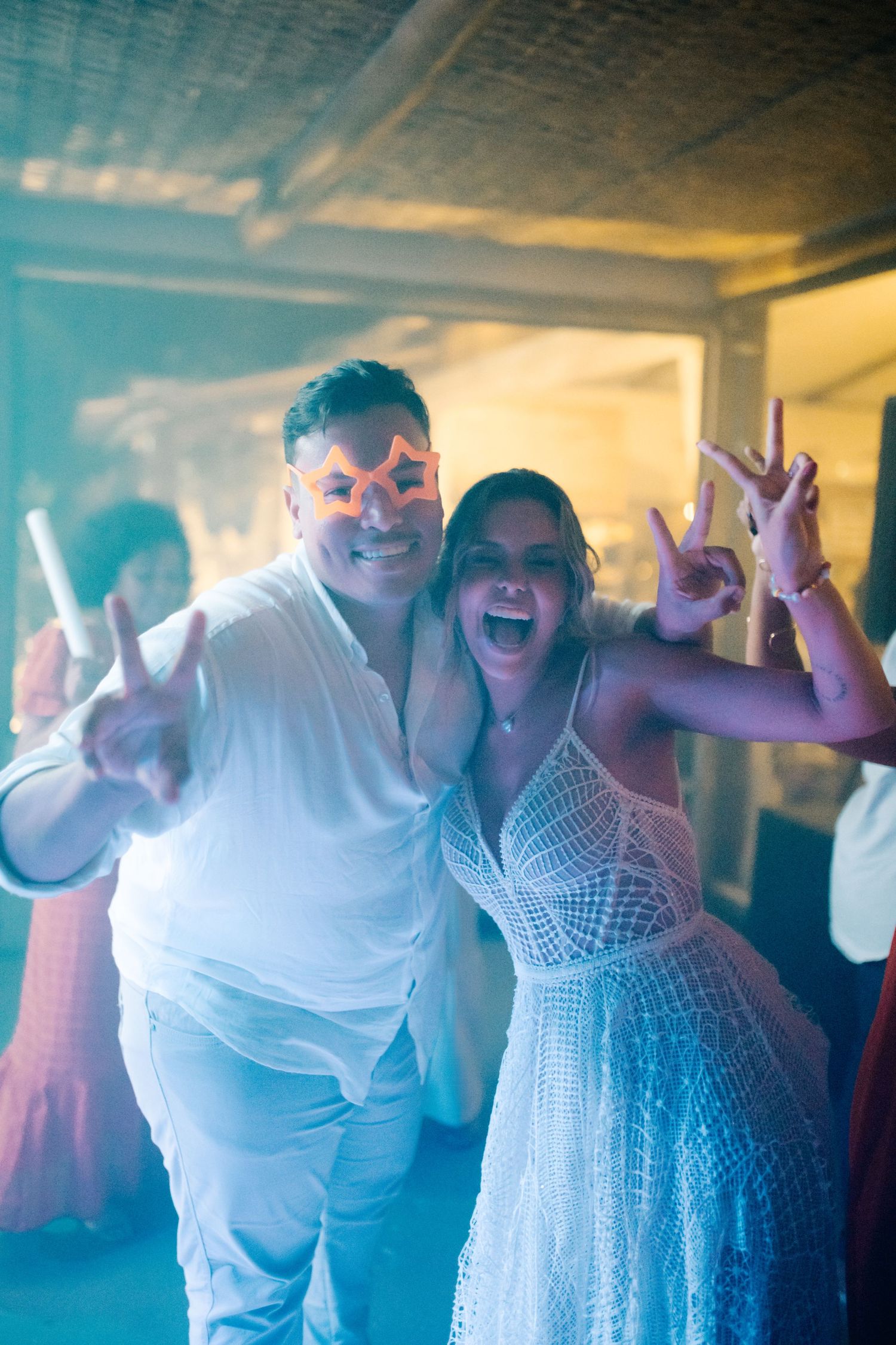 Party guests in white attire celebrate enthusiastically under colorful lighting.